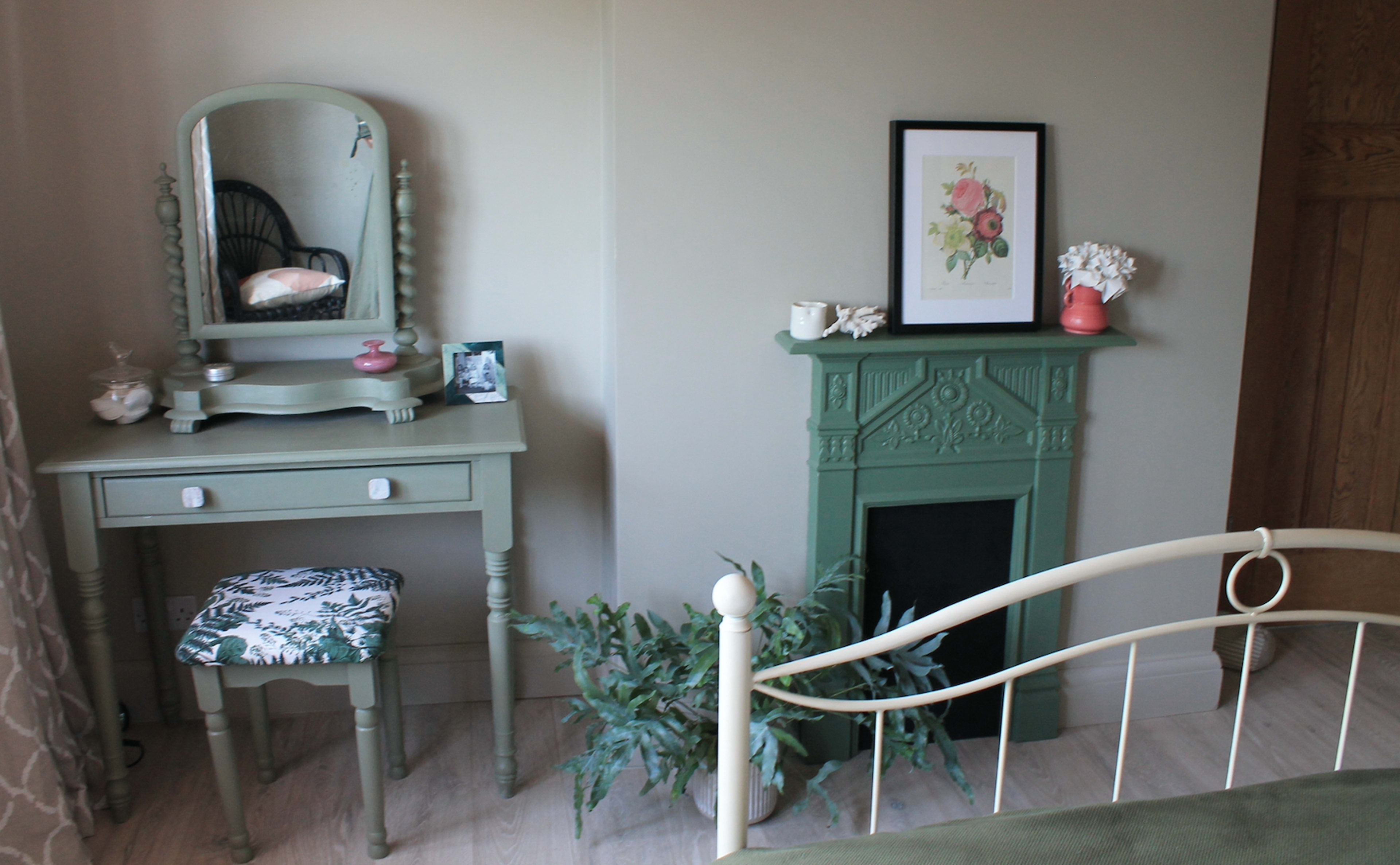 A small bedroom corner featuring a green vanity table with a mirror, a decorative framed flower print, and a matching green fireplace with a potted plant beside it.
