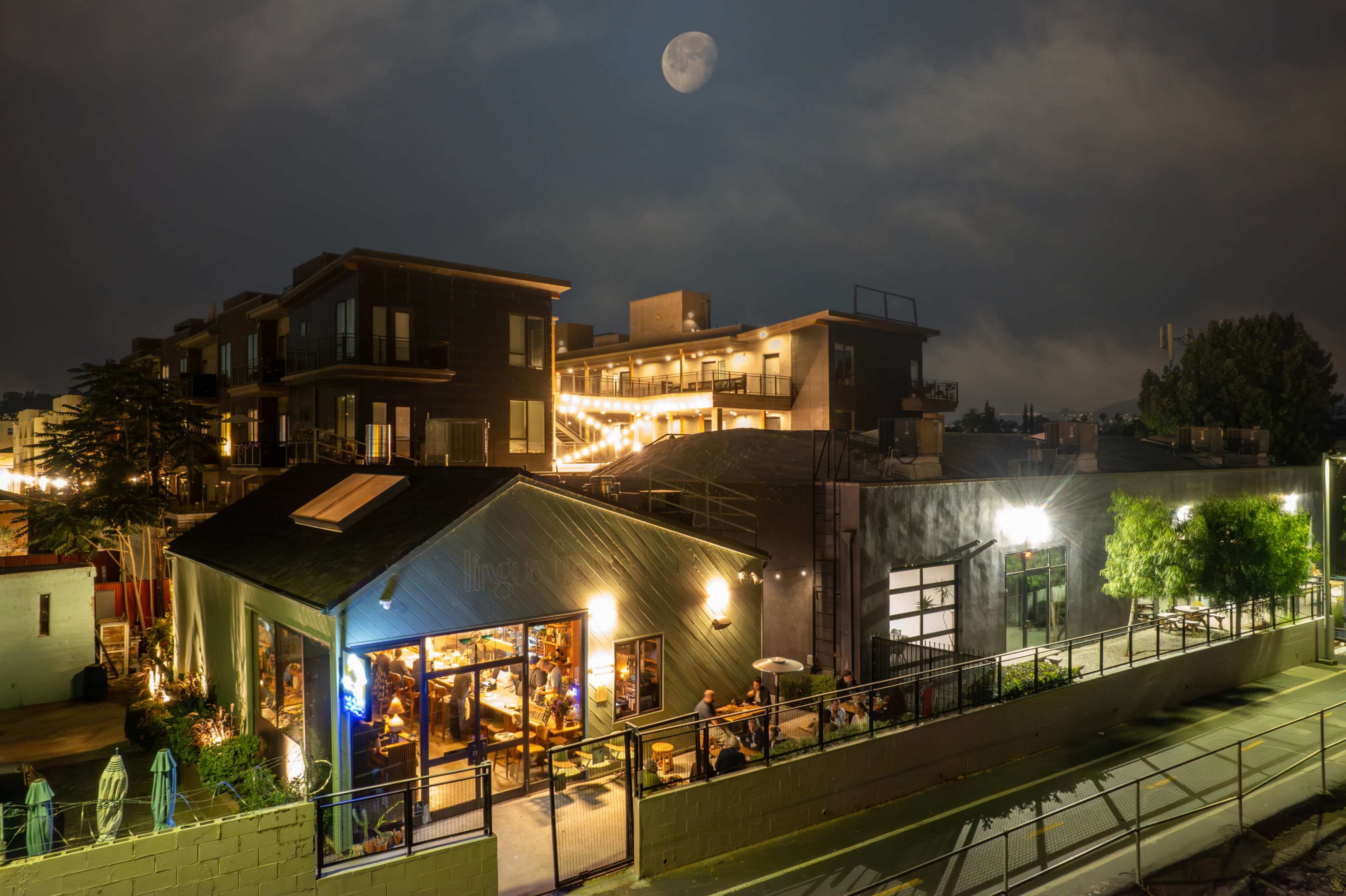 The scene shows a lively outdoor dining area of a restaurant illuminated at night, with a full moon visible in the sky above.