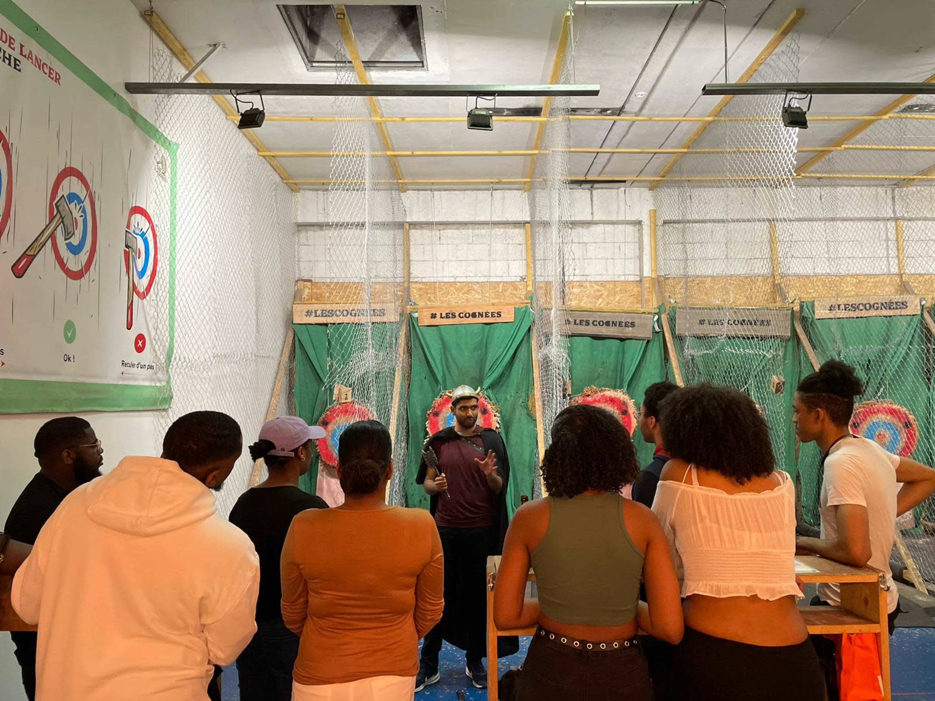 A group of people listens to a speaker in an indoor axe-throwing venue surrounded by targets and safety nets.