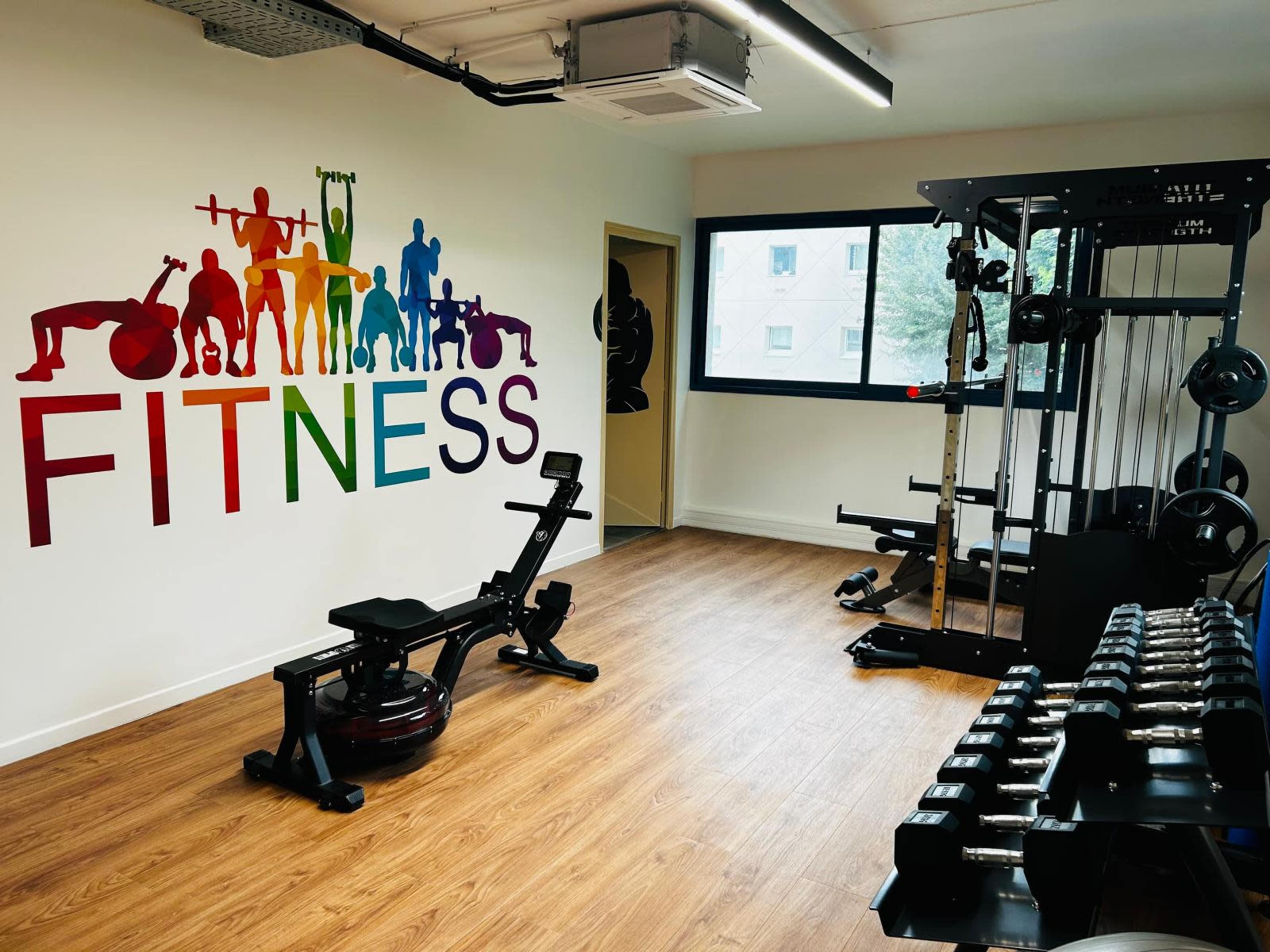 The image shows a fitness room featuring a colorful "FITNESS" mural, a workout bench, weightlifting equipment, a rowing machine, and a set of dumbbells.