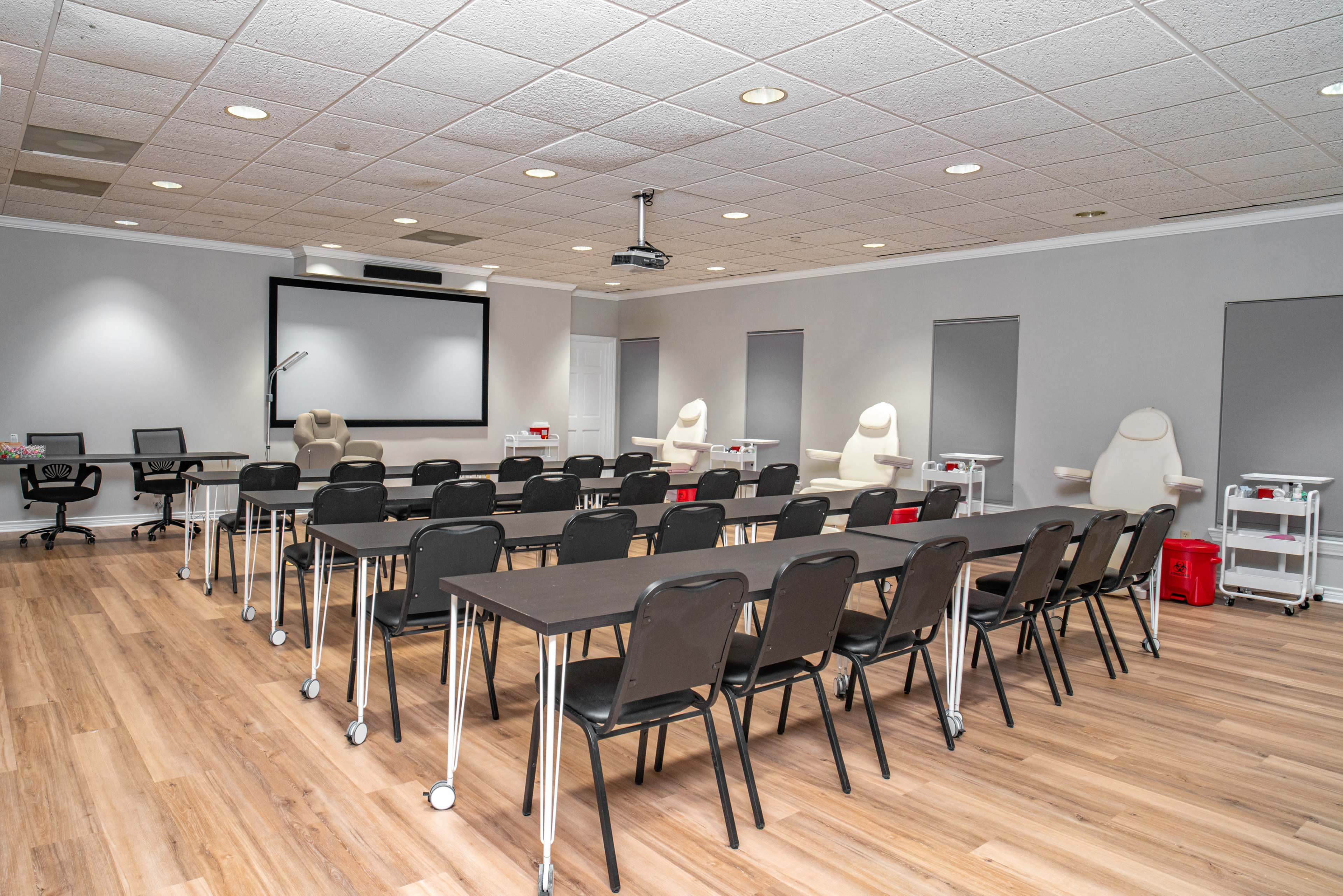 A training room features rows of black chairs and long tables facing a projection screen, with additional equipment and supplies in the background.