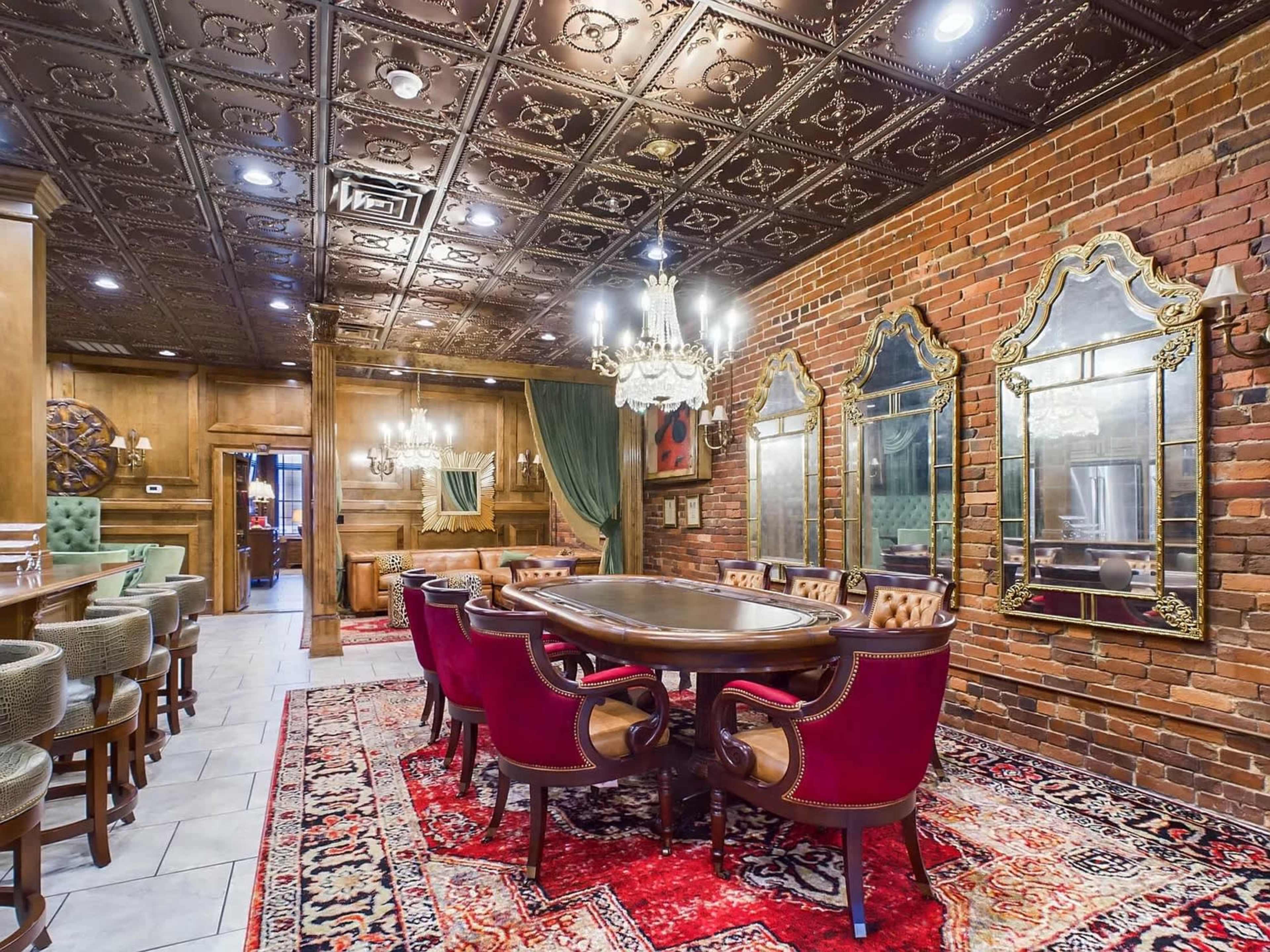 The image shows a dining room with a round table surrounded by red chairs, brick walls, and ornate mirrors, under an ornate ceiling with chandeliers.