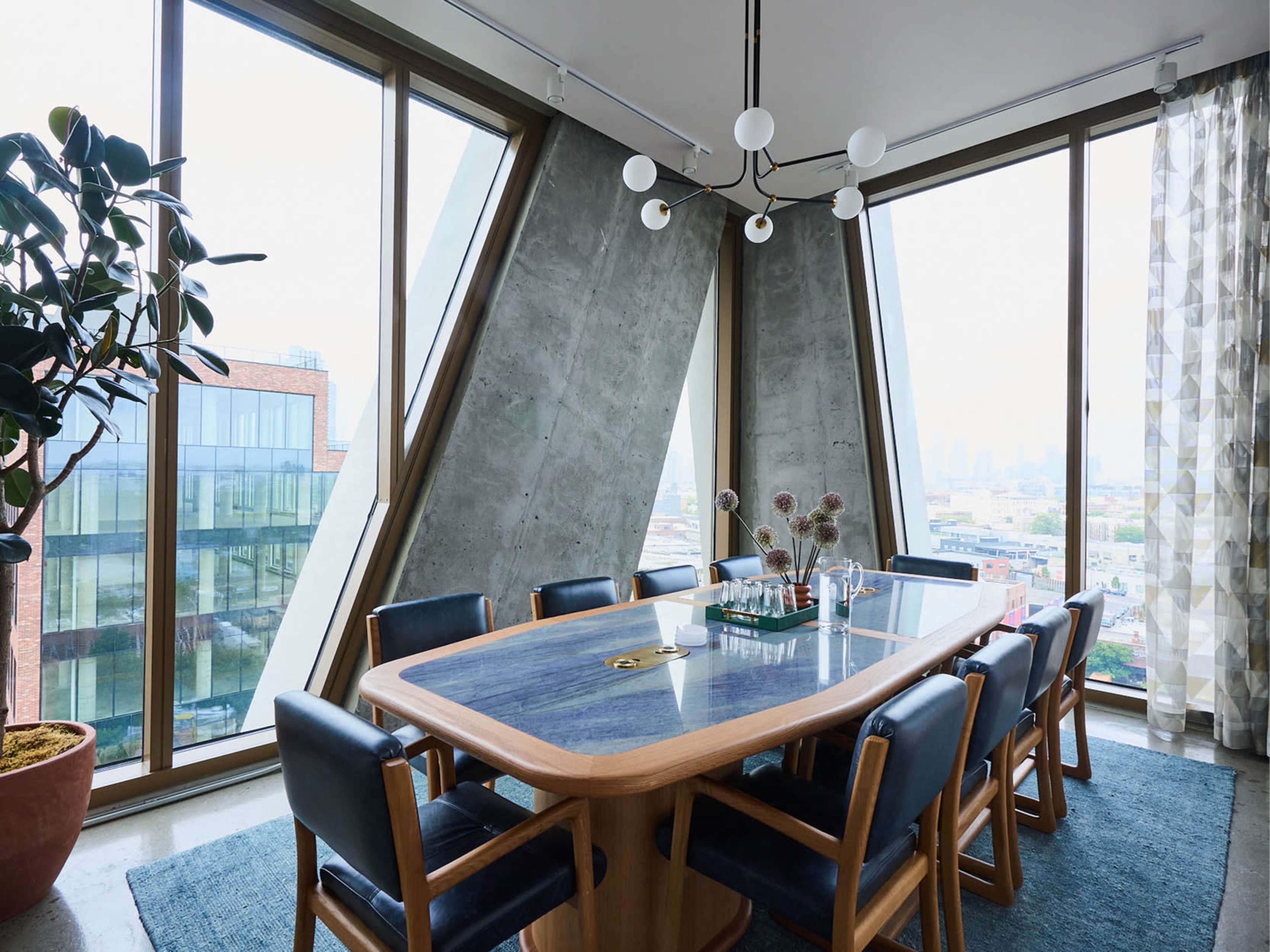 A modern dining room features a large wooden table surrounded by blue chairs, with large windows showcasing an urban view.