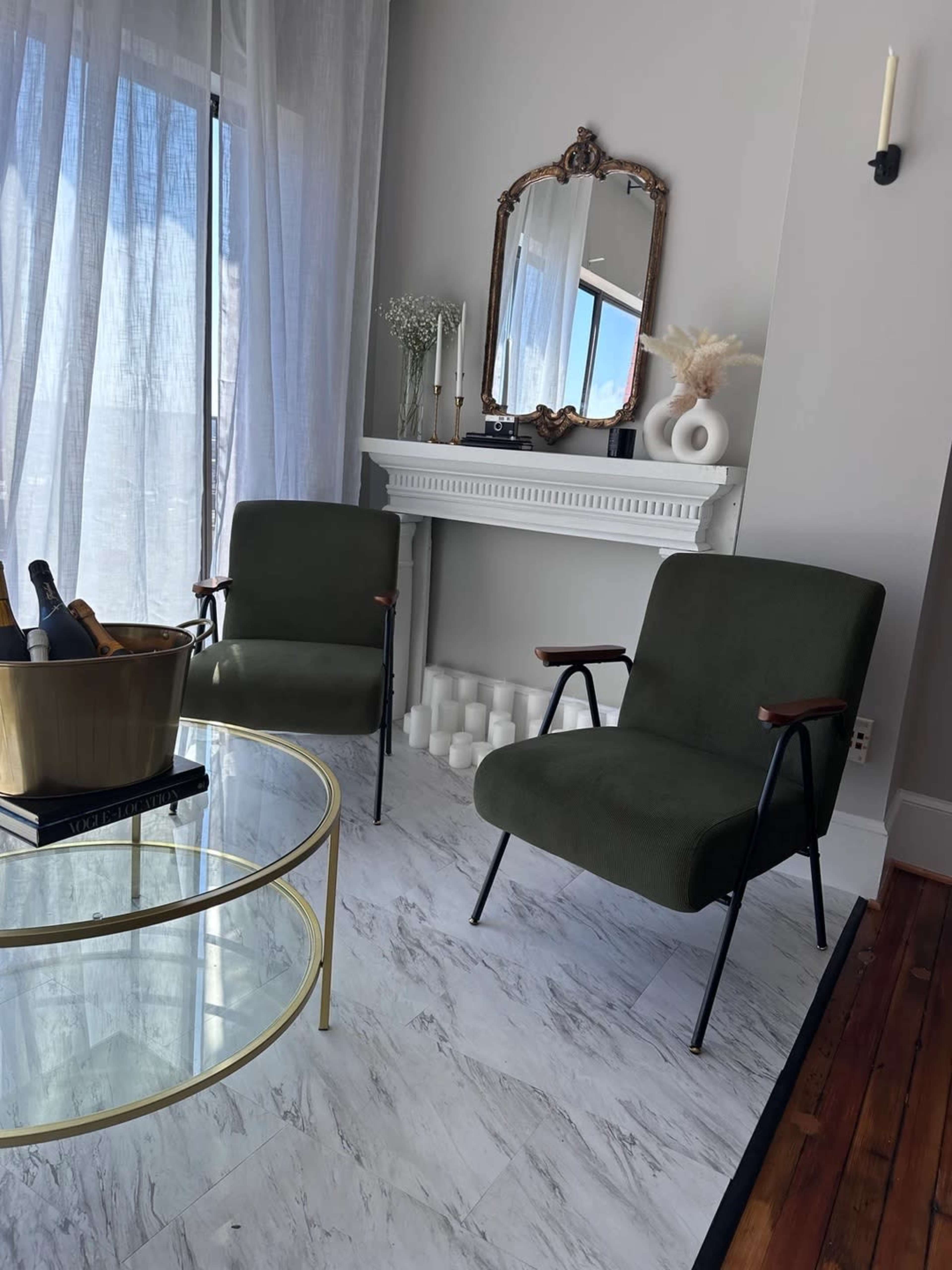 A modern living room with two green chairs facing a glass coffee table, near a fireplace with decorative items and soft natural light coming through sheer curtains.