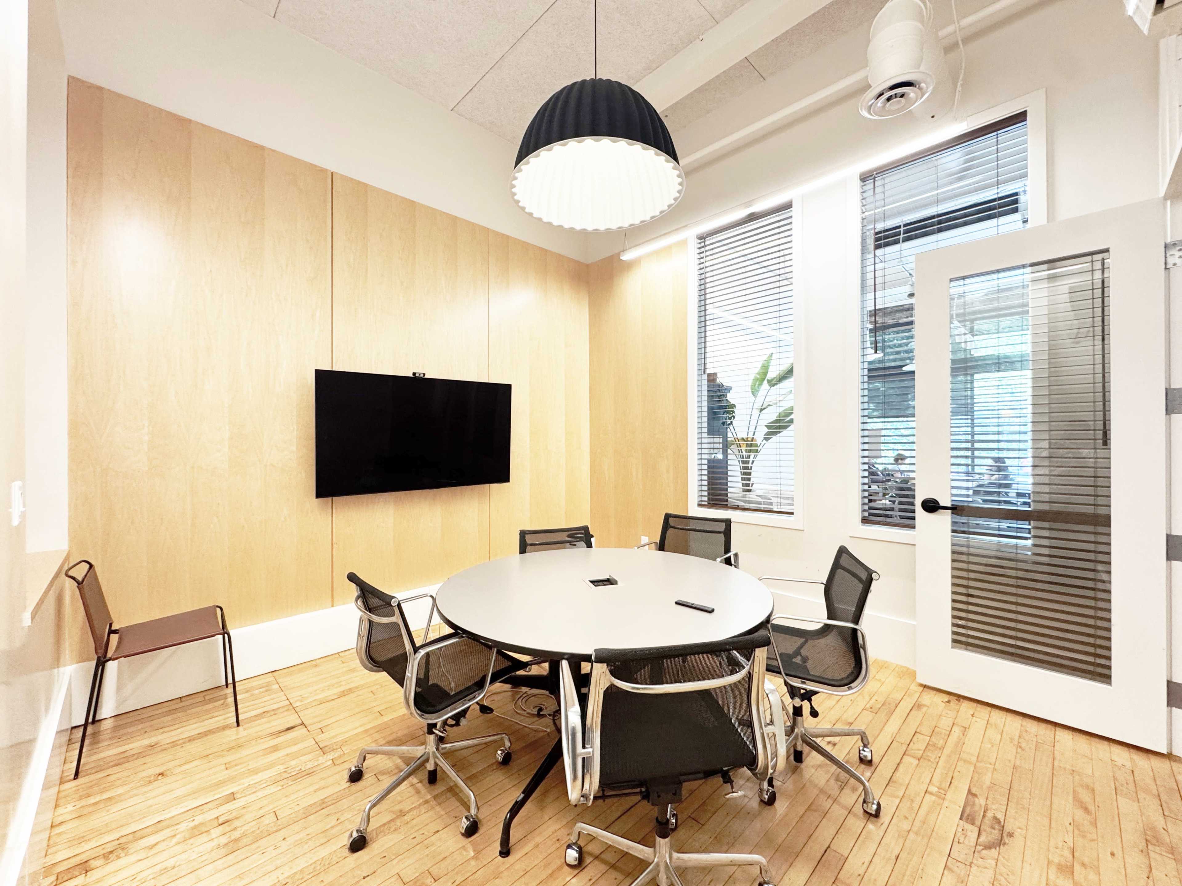A modern conference room features a round table surrounded by six chairs, a large television mounted on the wall, and natural light from windows with blinds.