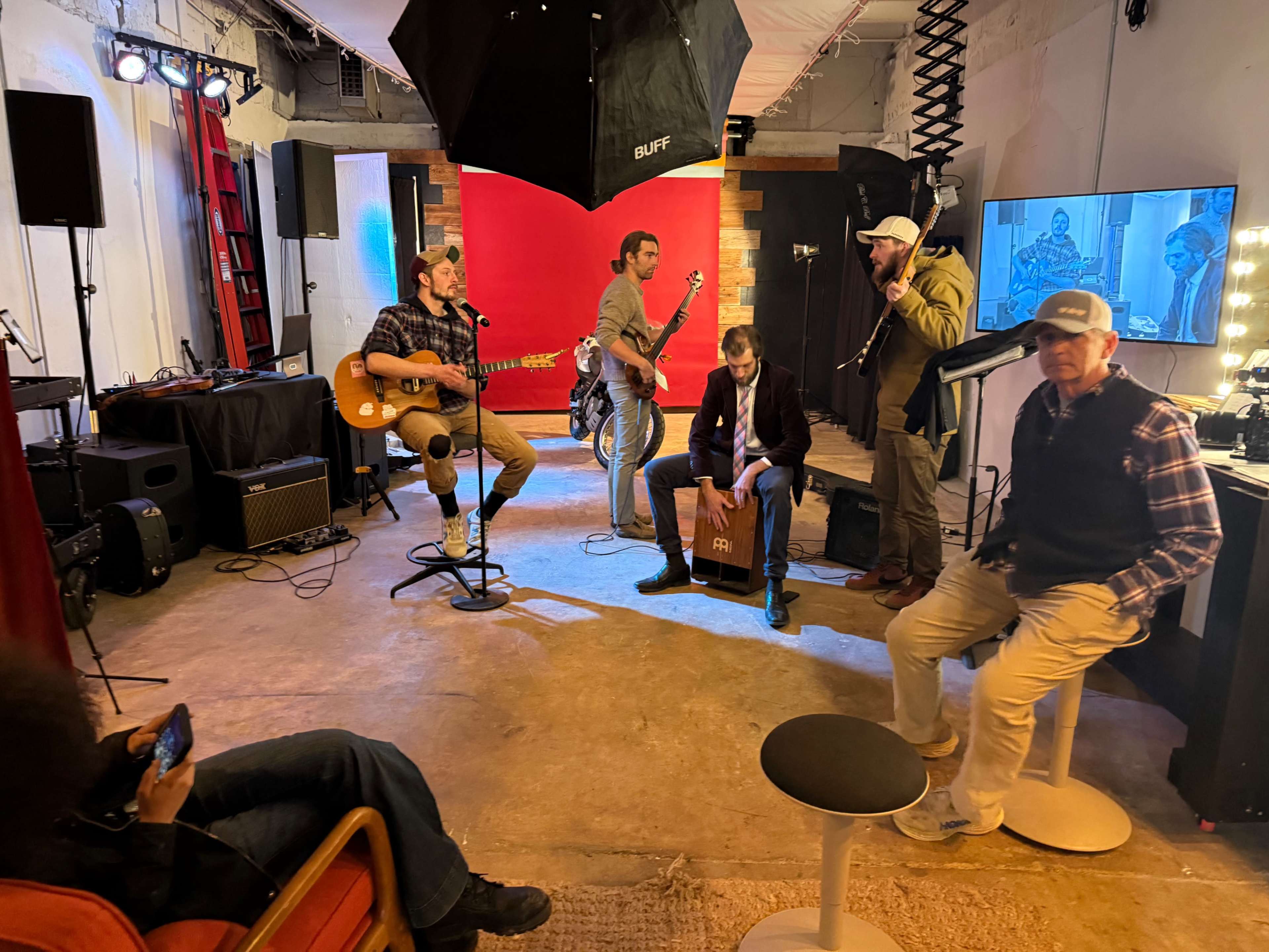 A group of five musicians performs in a studio setting equipped with musical instruments, a microphone, and a screen displaying images.