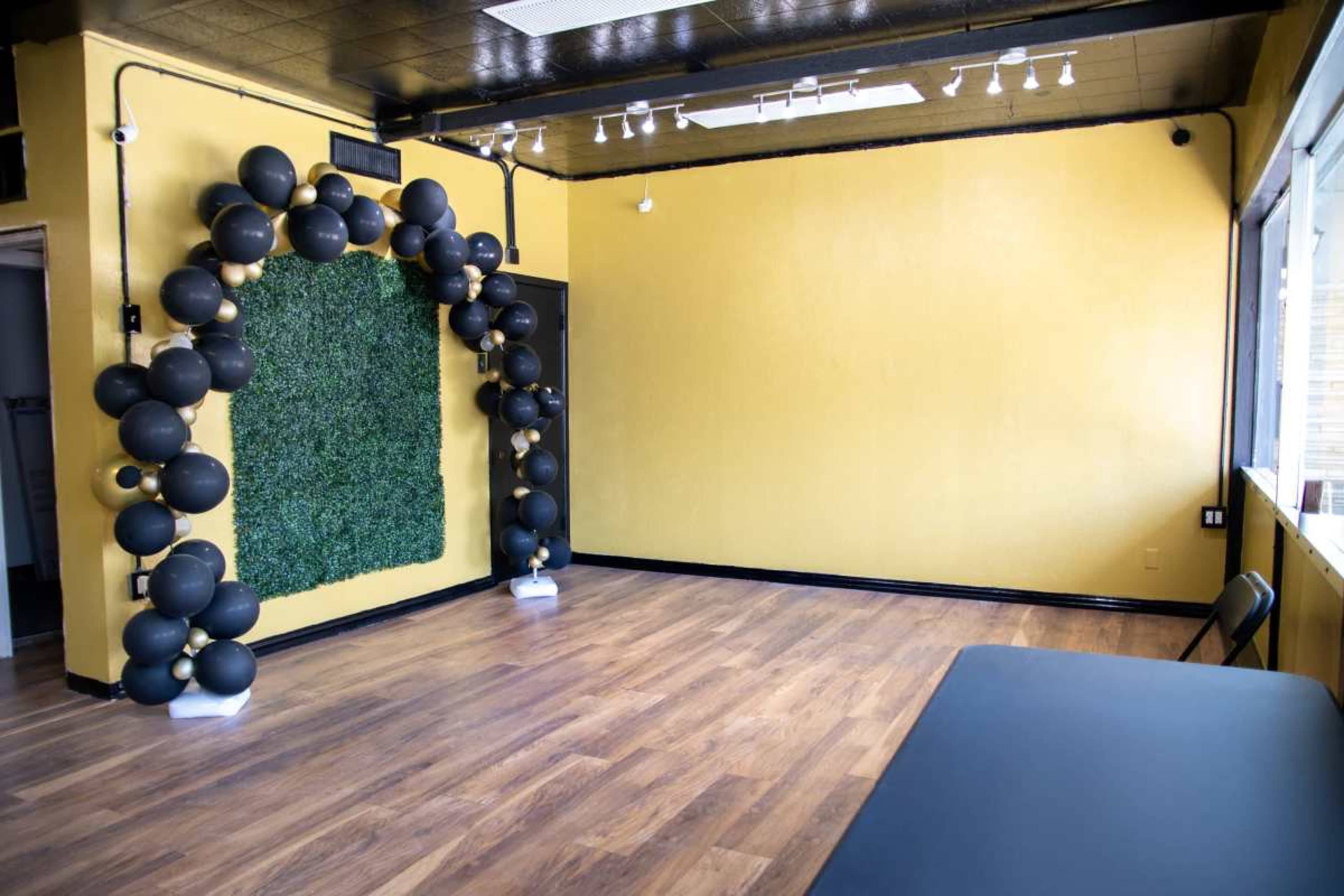 An empty room with yellow walls, wooden flooring, and a decorative arch made of black balloons next to a wall covered in green foliage.