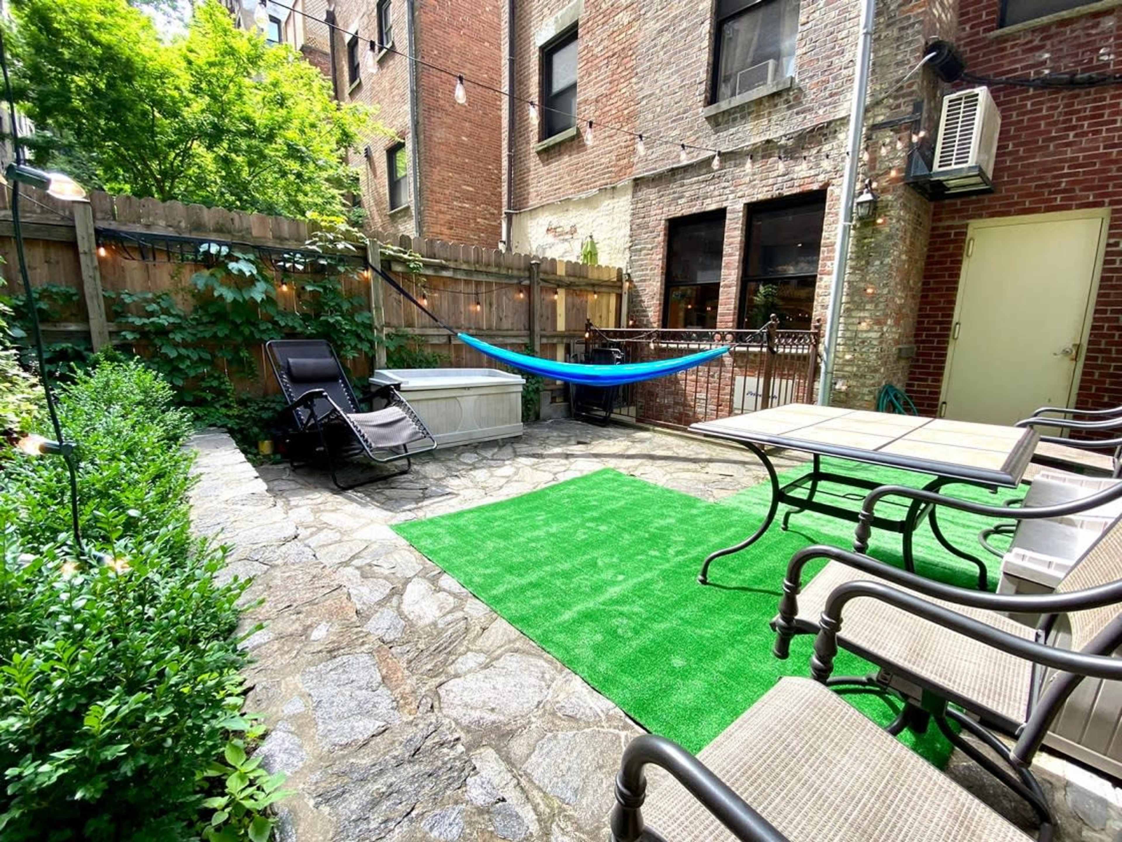 A small outdoor patio area with stone flooring, a green rug, a hammock, lounge chairs, and a table surrounded by greenery and brick walls.