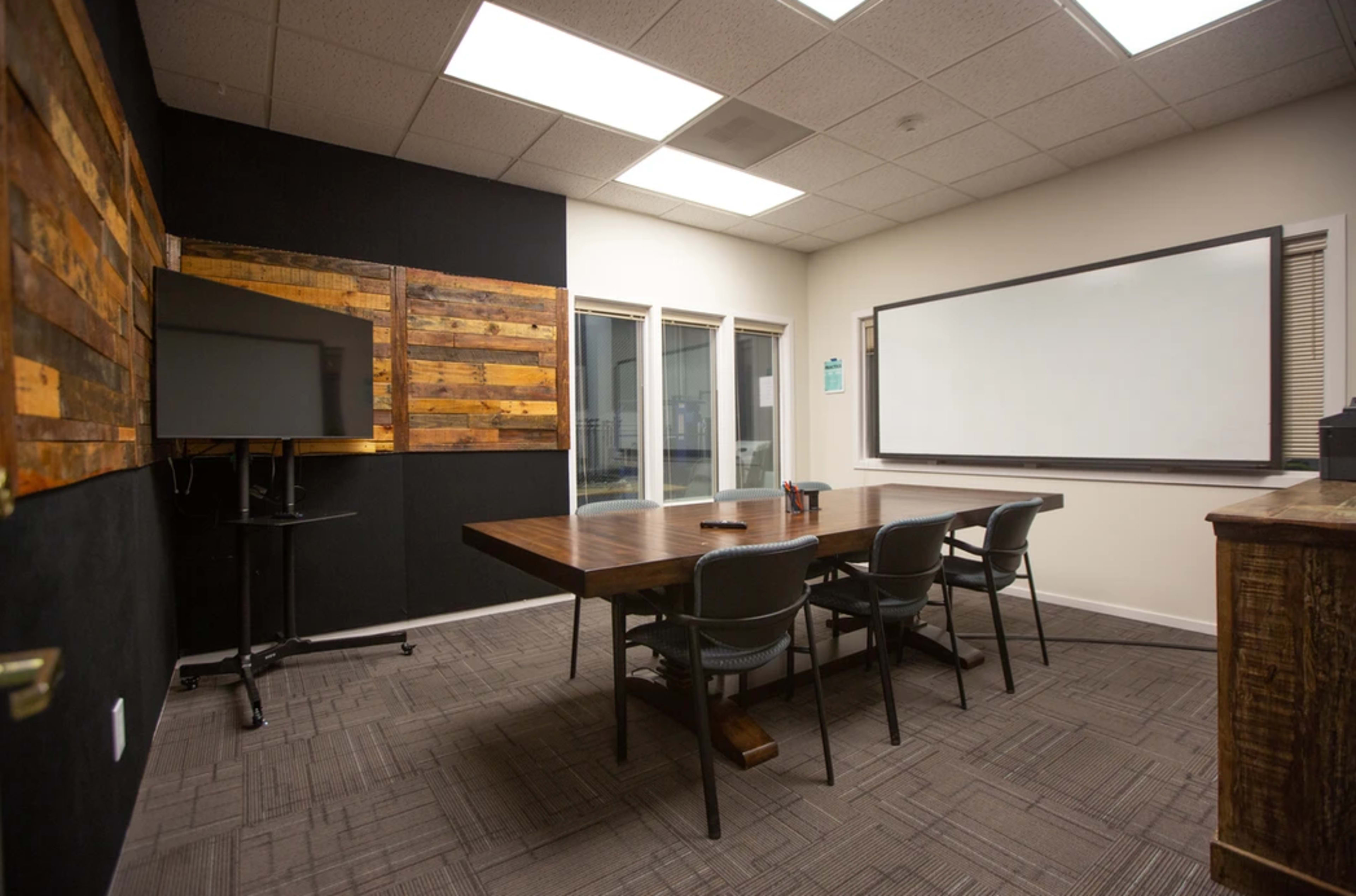 The image shows a conference room with a large wooden table, four chairs, a mounted screen, and a whiteboard on the wall.