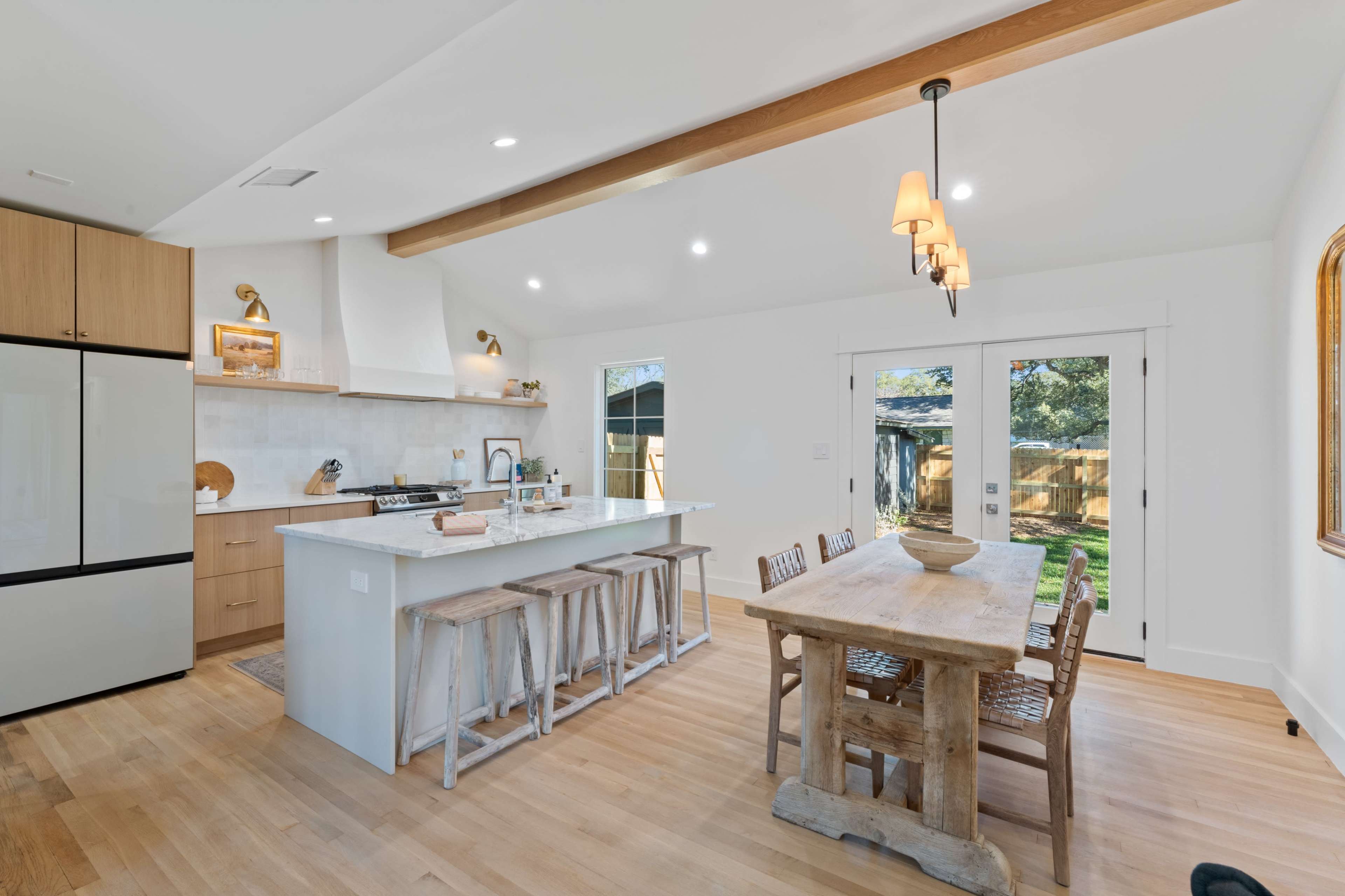 A modern kitchen features a large island with seating, wooden beams on the ceiling, and a rustic dining table under pendant lights.
