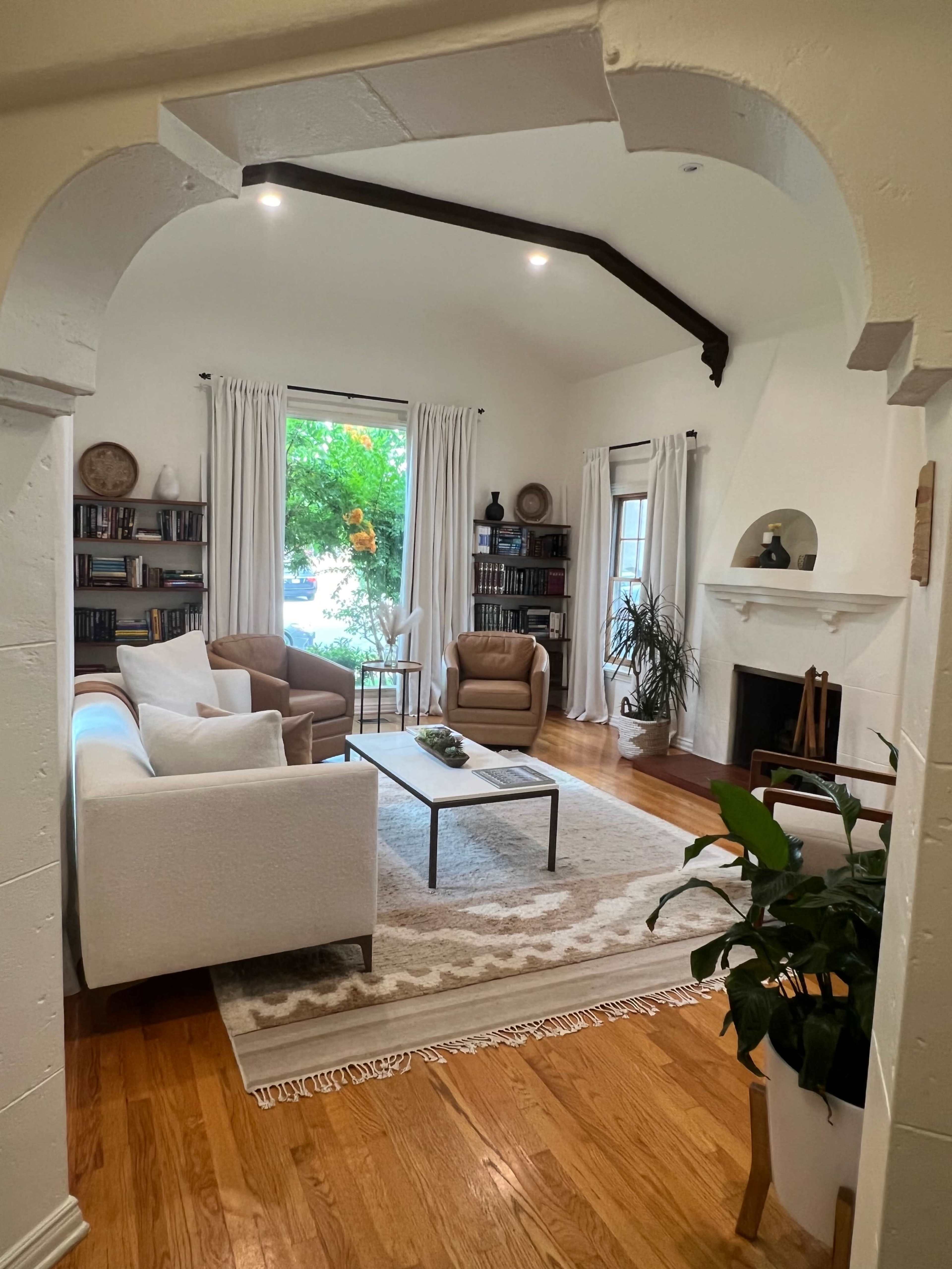 A cozy living room features a pair of armchairs and a white couch arranged around a coffee table on a textured rug, with bookshelves and a fireplace in the background.