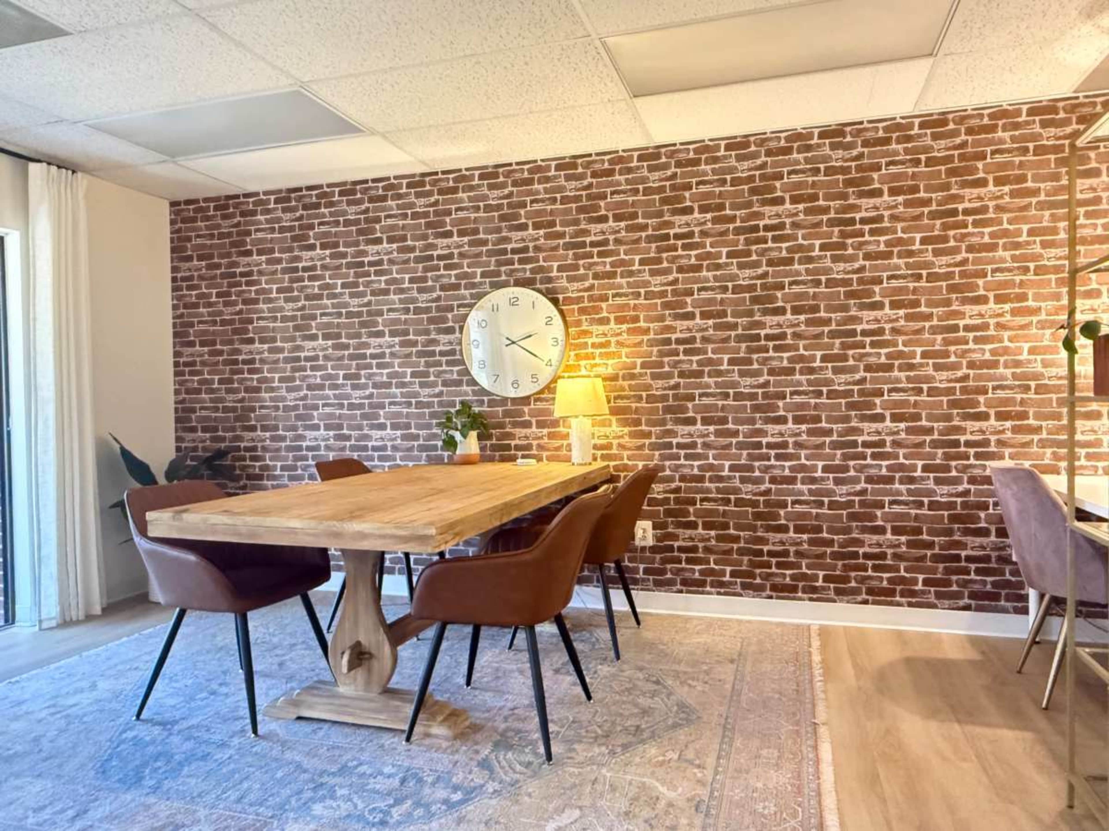 A dining area features a wooden table surrounded by brown chairs, with a brick-patterned wall and a large clock.