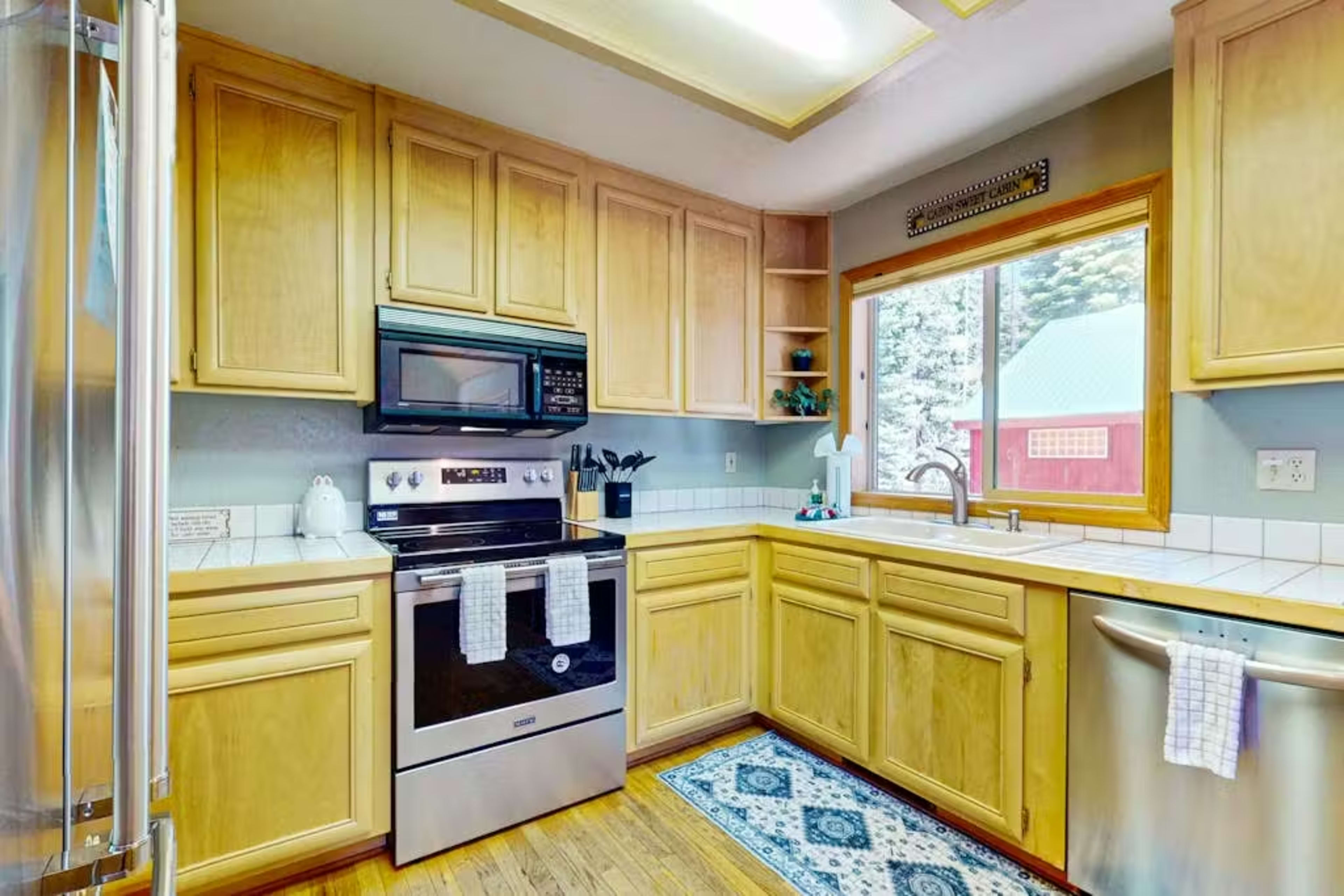 The image shows a compact kitchen with wooden cabinetry, a stainless steel refrigerator, a black oven and microwave, a stainless steel sink, and a window providing a view of a red structure outside.