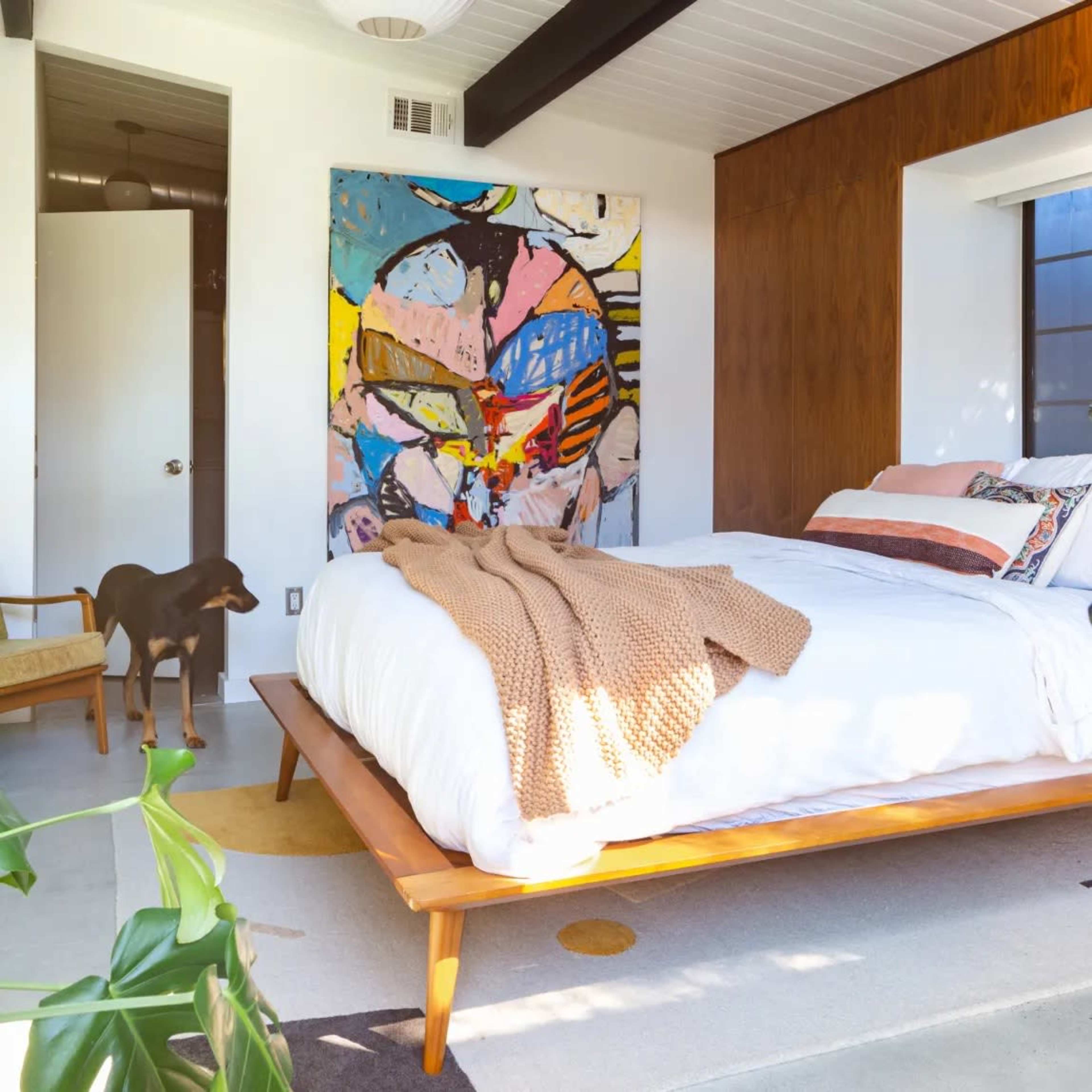 A modern bedroom features a bed with white linens and a colorful abstract painting on the wall, with a dog standing near a chair.