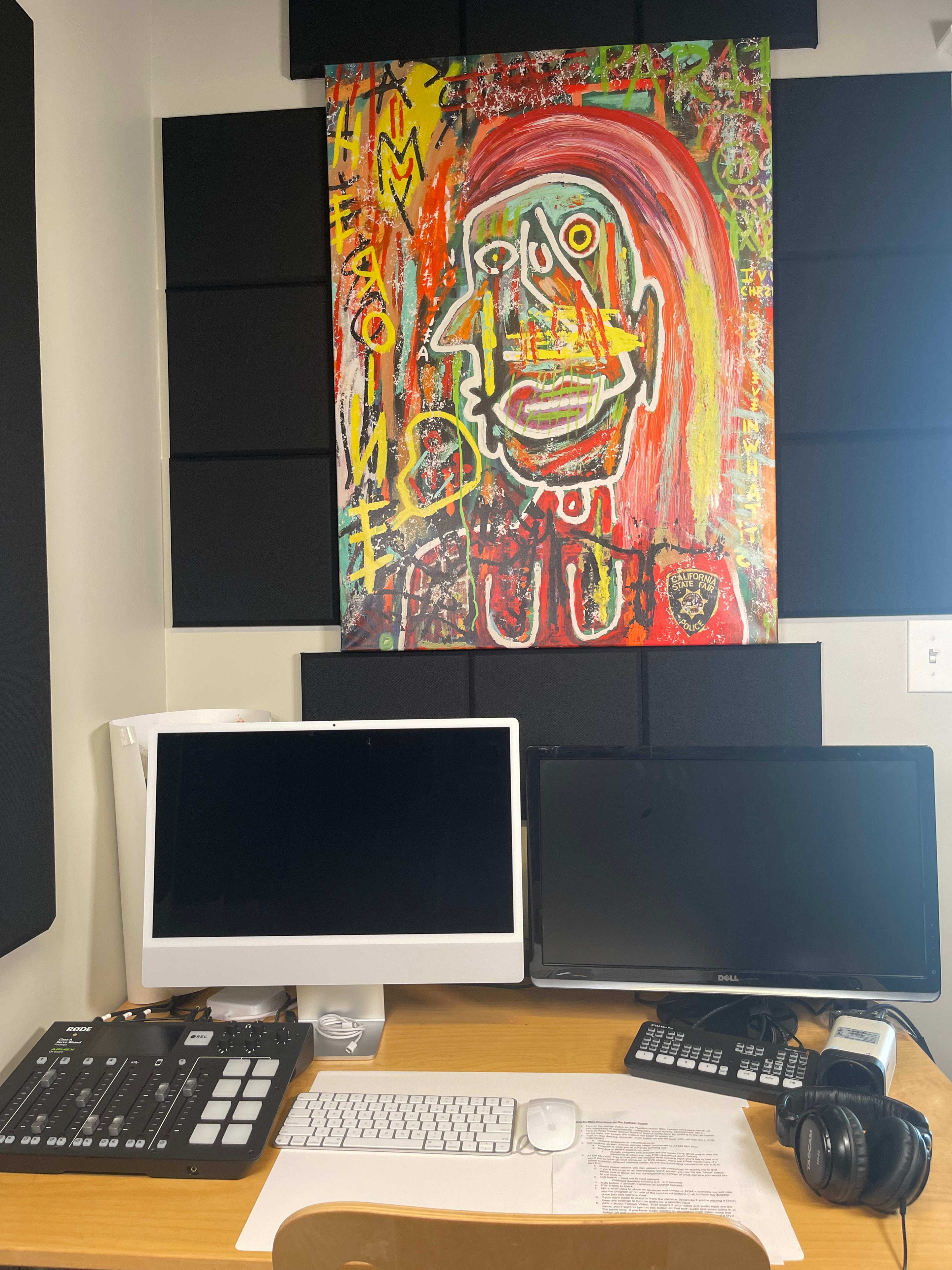 A workspace featuring two computer monitors, a MIDI pad, a telephone, and a colorful abstract painting on the wall.
