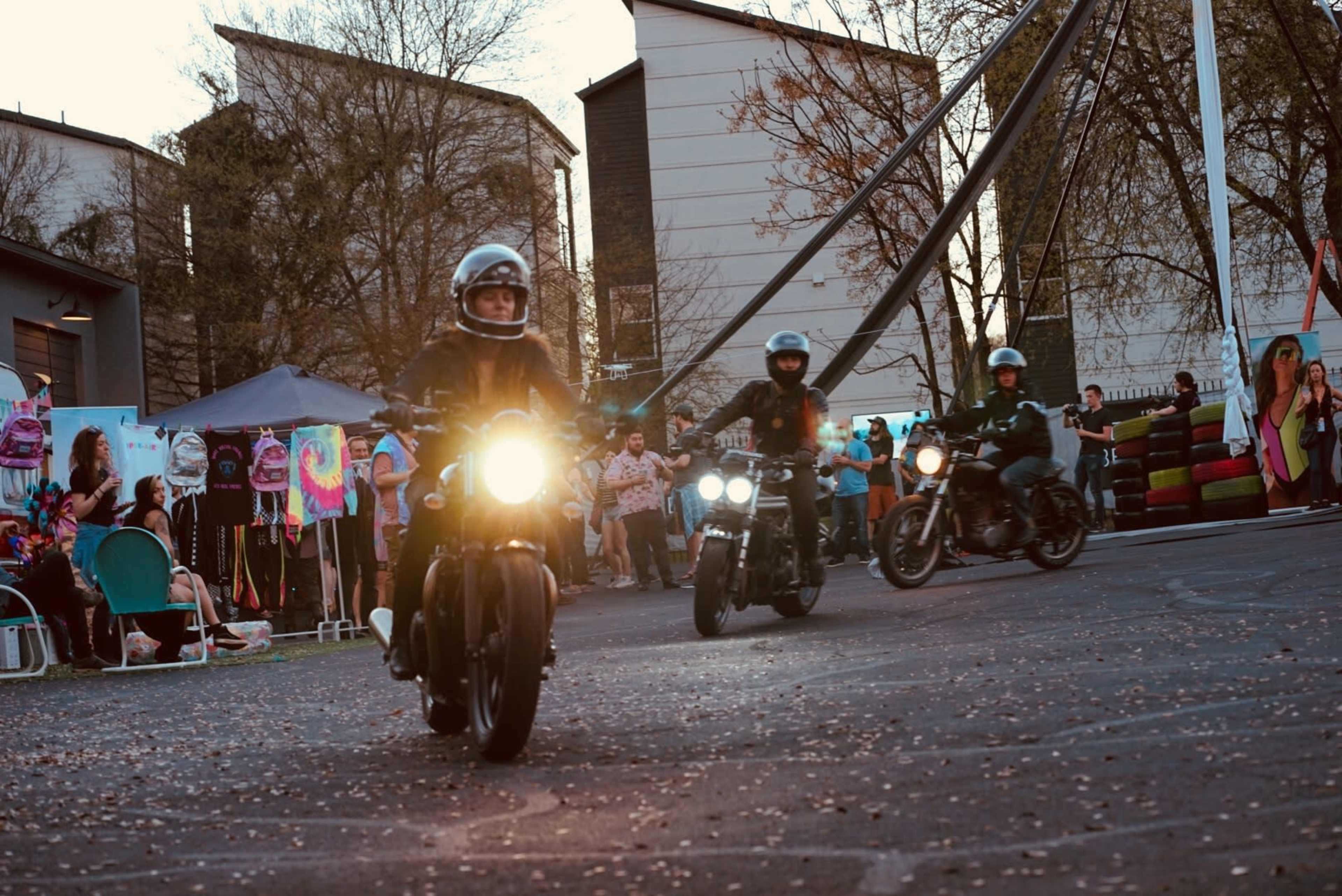 Three motorcyclists ride through a festival scene with spectators and vendor tents visible in the background.