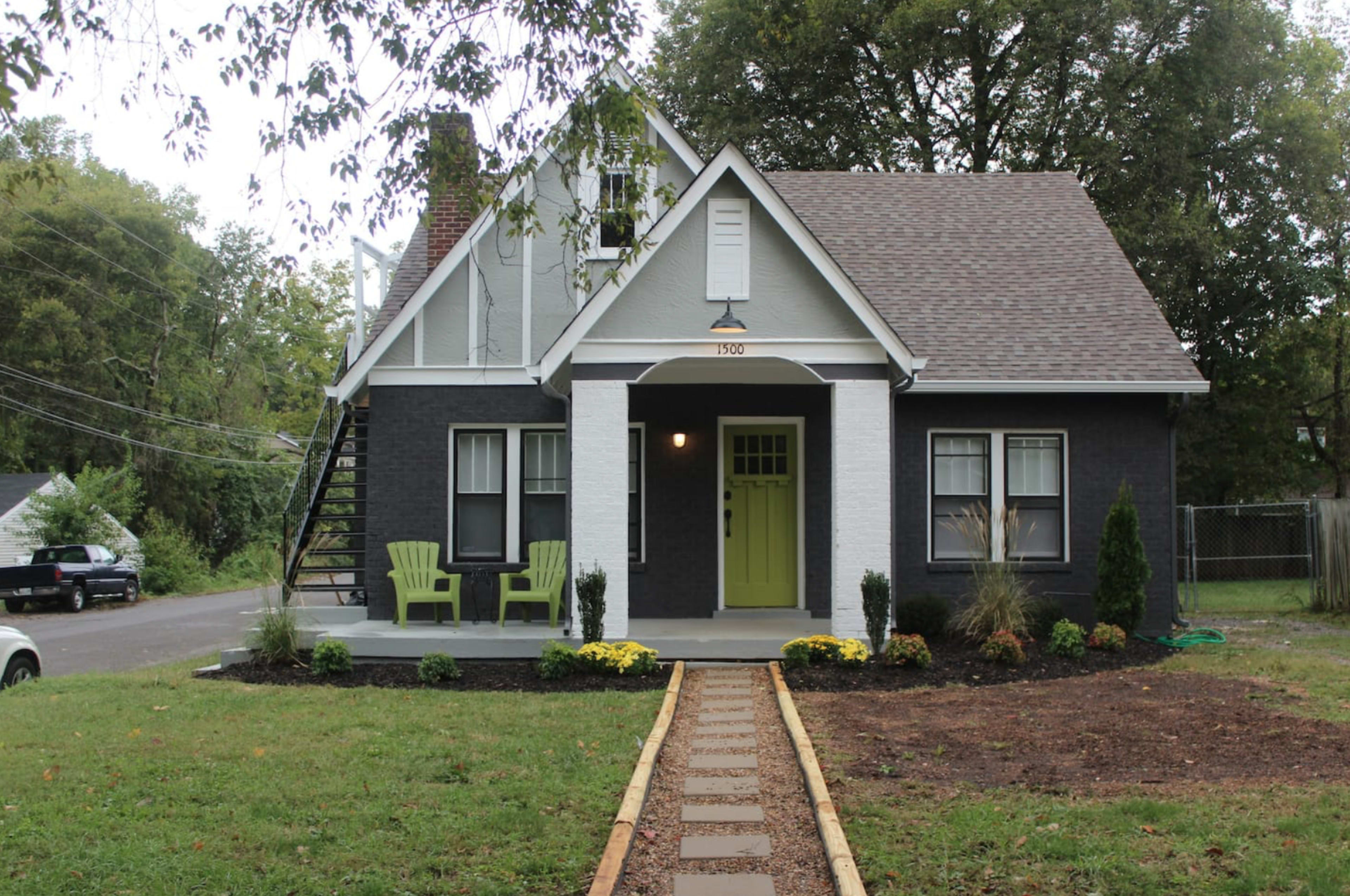 A two-story house with dark siding and a white trim features a green front door and a pathway lined with flowers and trees.
