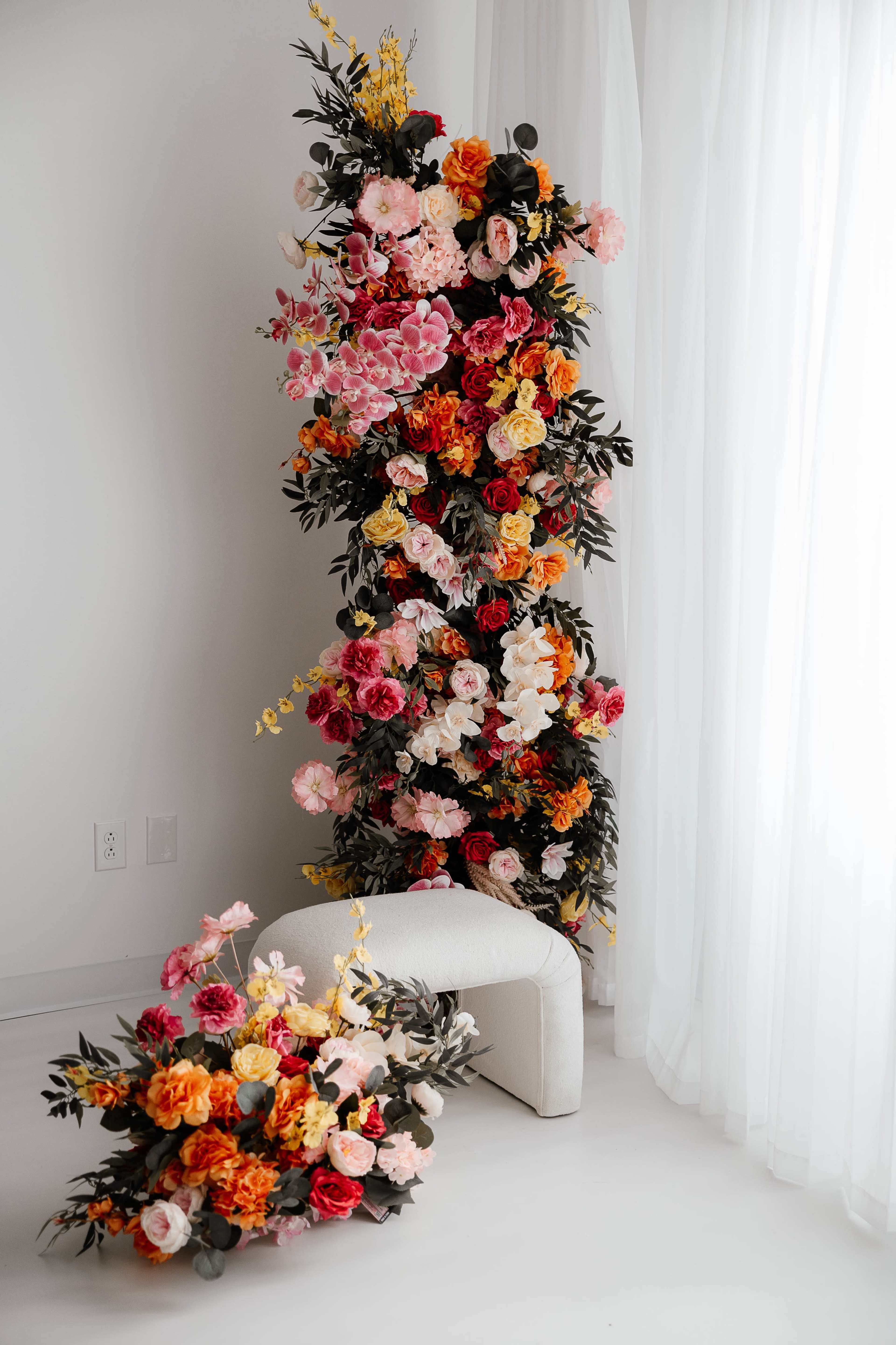A vertical floral arrangement composed of various colored flowers is positioned next to a small, white, upholstered bench against a backdrop of sheer curtains.