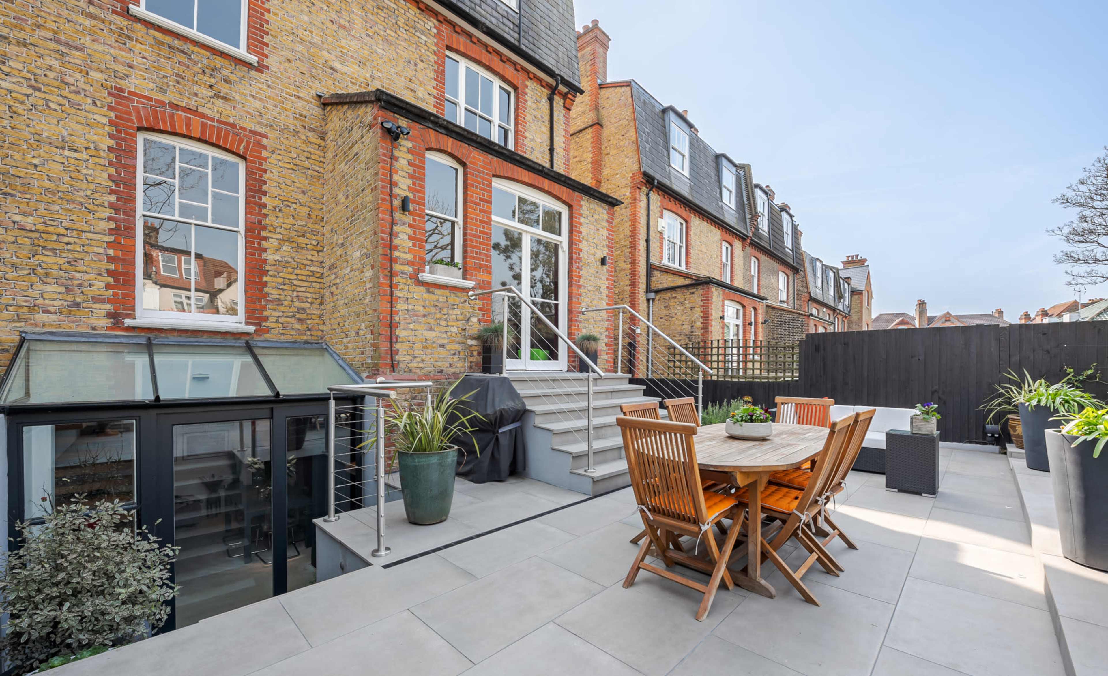 The image shows a backyard patio with a wooden dining table and chairs, leading up to a brick building with multiple windows.