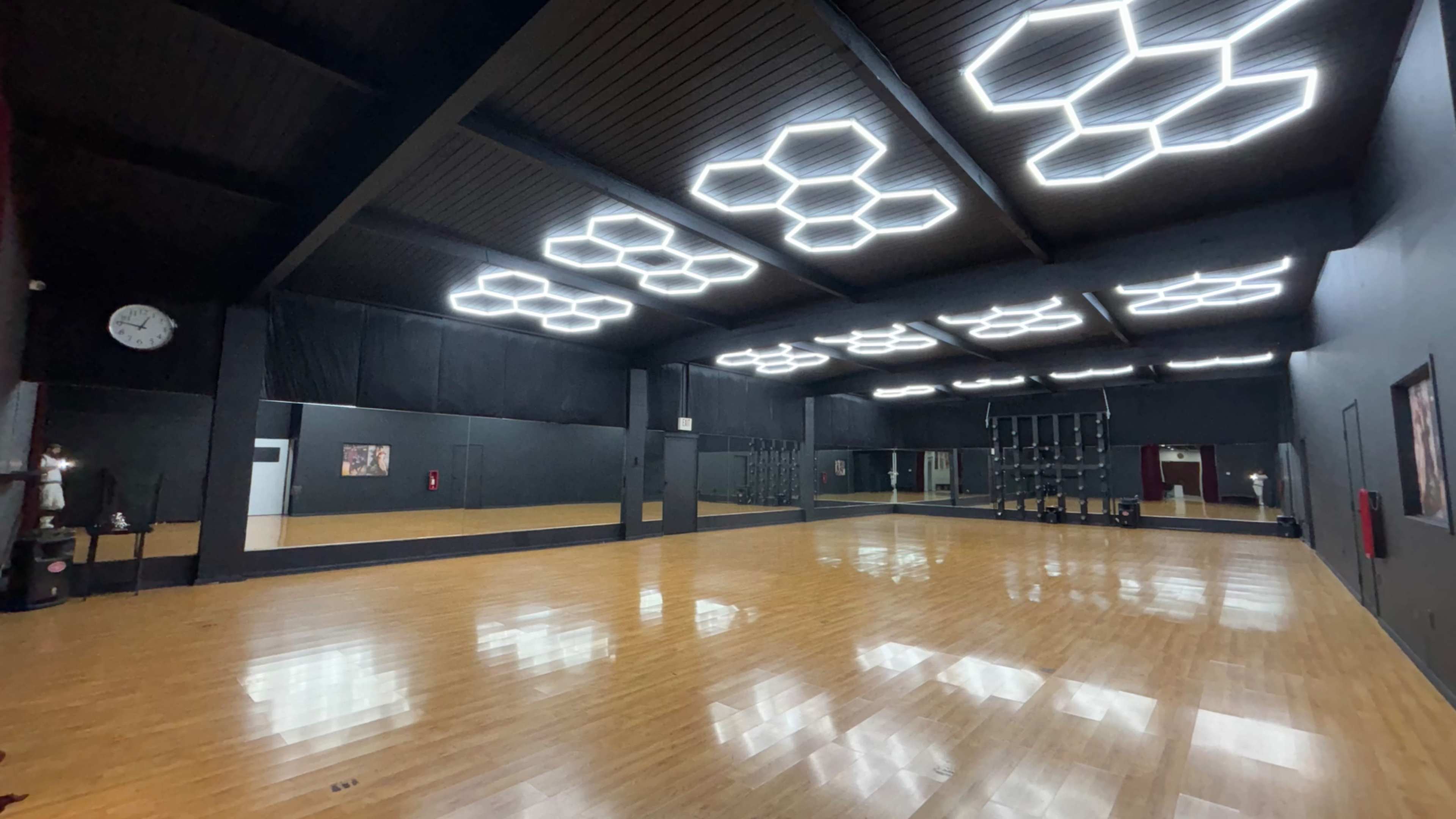 The image shows a spacious dance studio with hexagonal ceiling lights, a polished wooden floor, and mirrored walls.