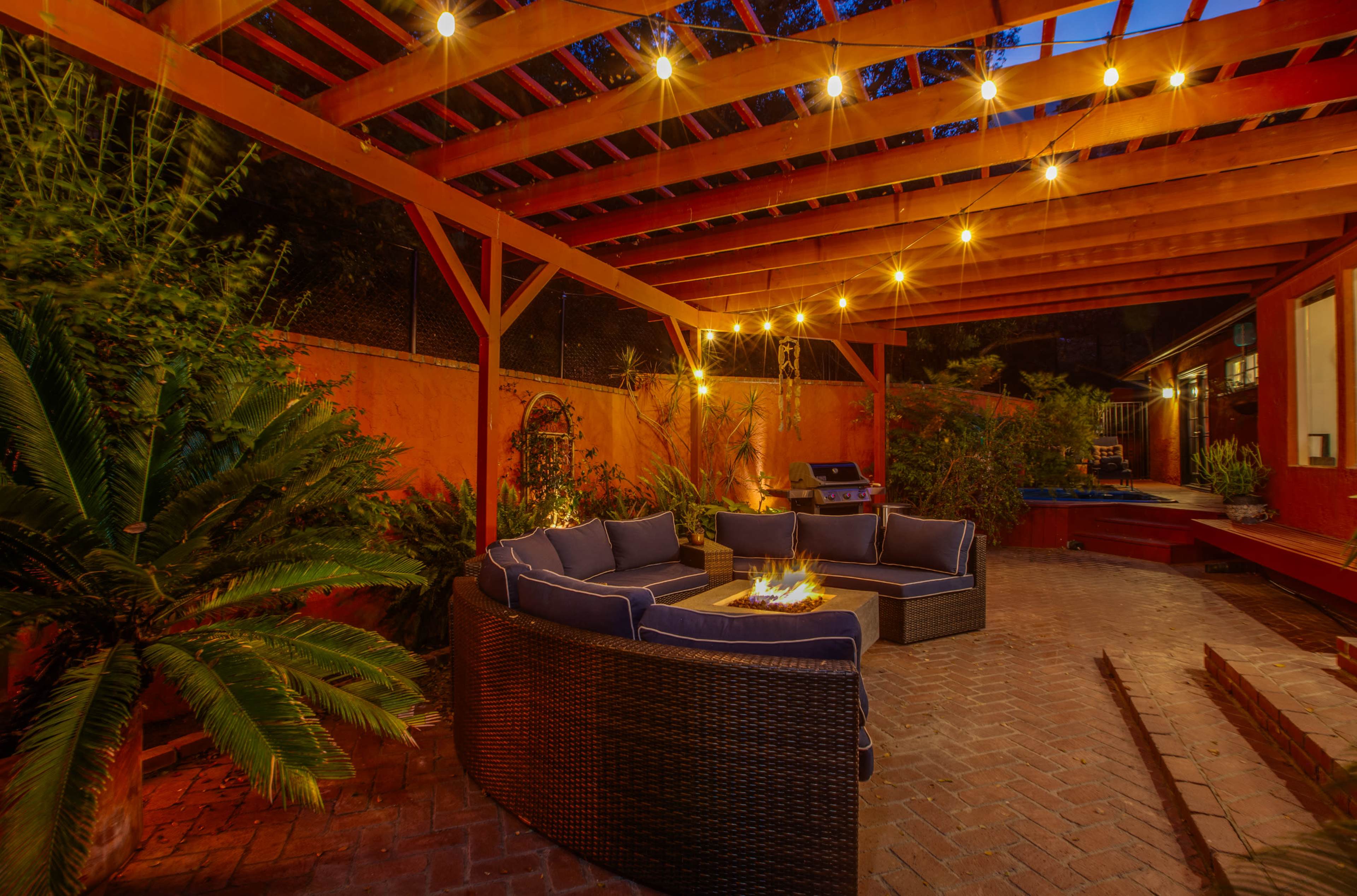 A cozy outdoor seating area features a circular arrangement of dark wicker chairs around a fire pit, with string lights illuminating the space under a wooden pergola.