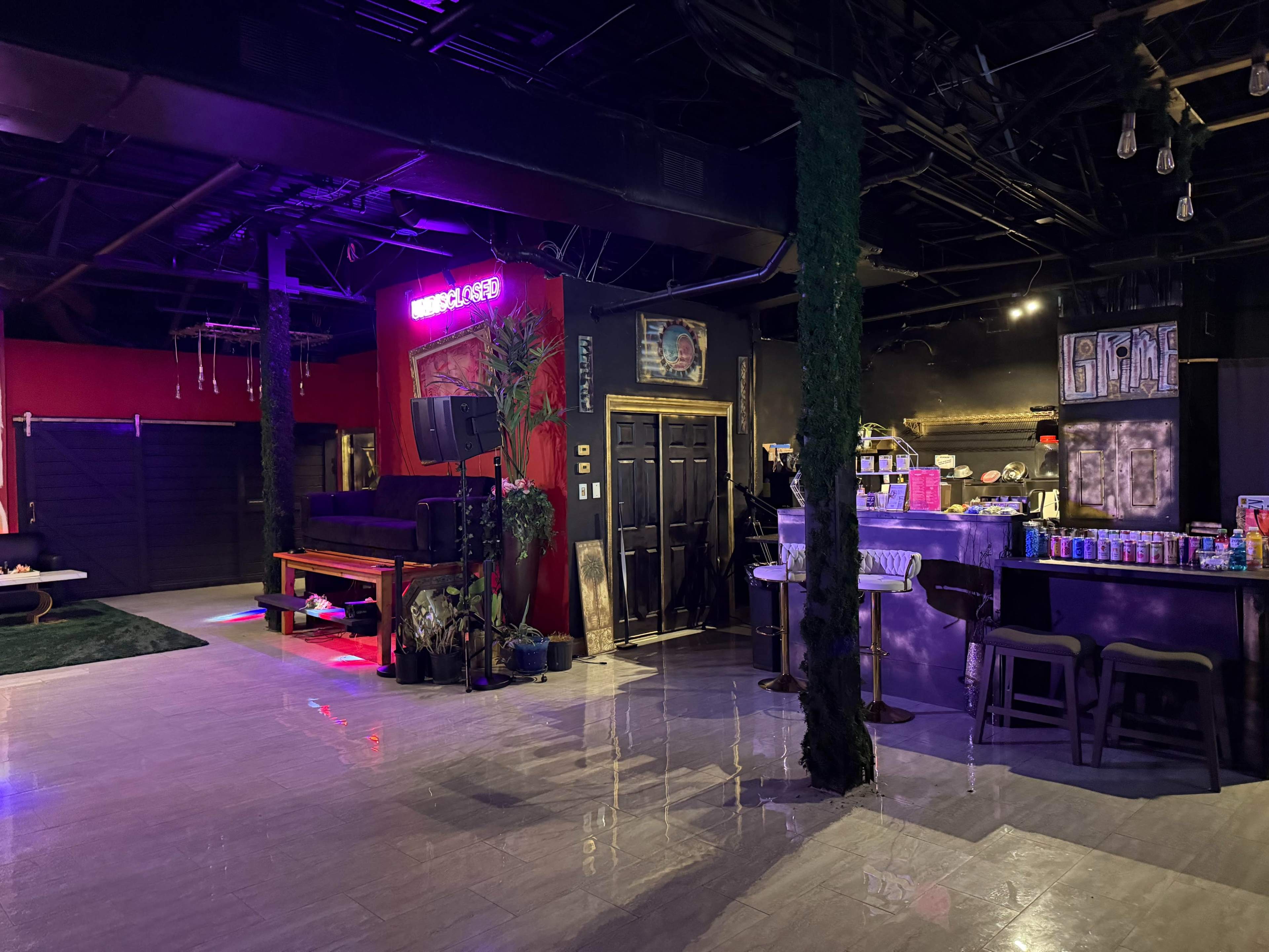 The image shows a dimly lit indoor space with a bar area on one side, a lounge area with seating, and neon lights illuminating the scene.