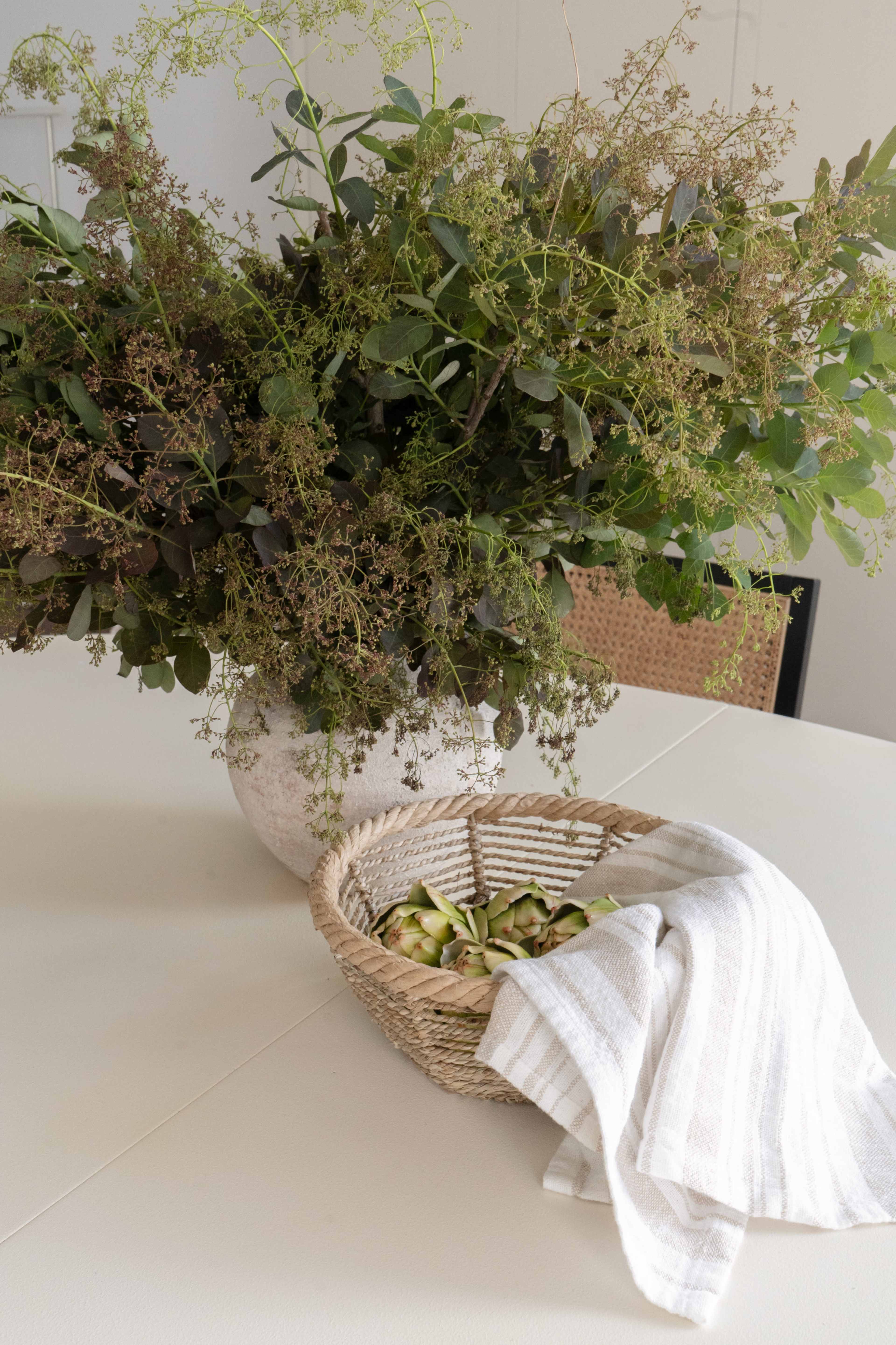 A large bouquet of greenery sits in a textured vase next to a woven basket containing sliced vegetables and a striped towel on a white table.