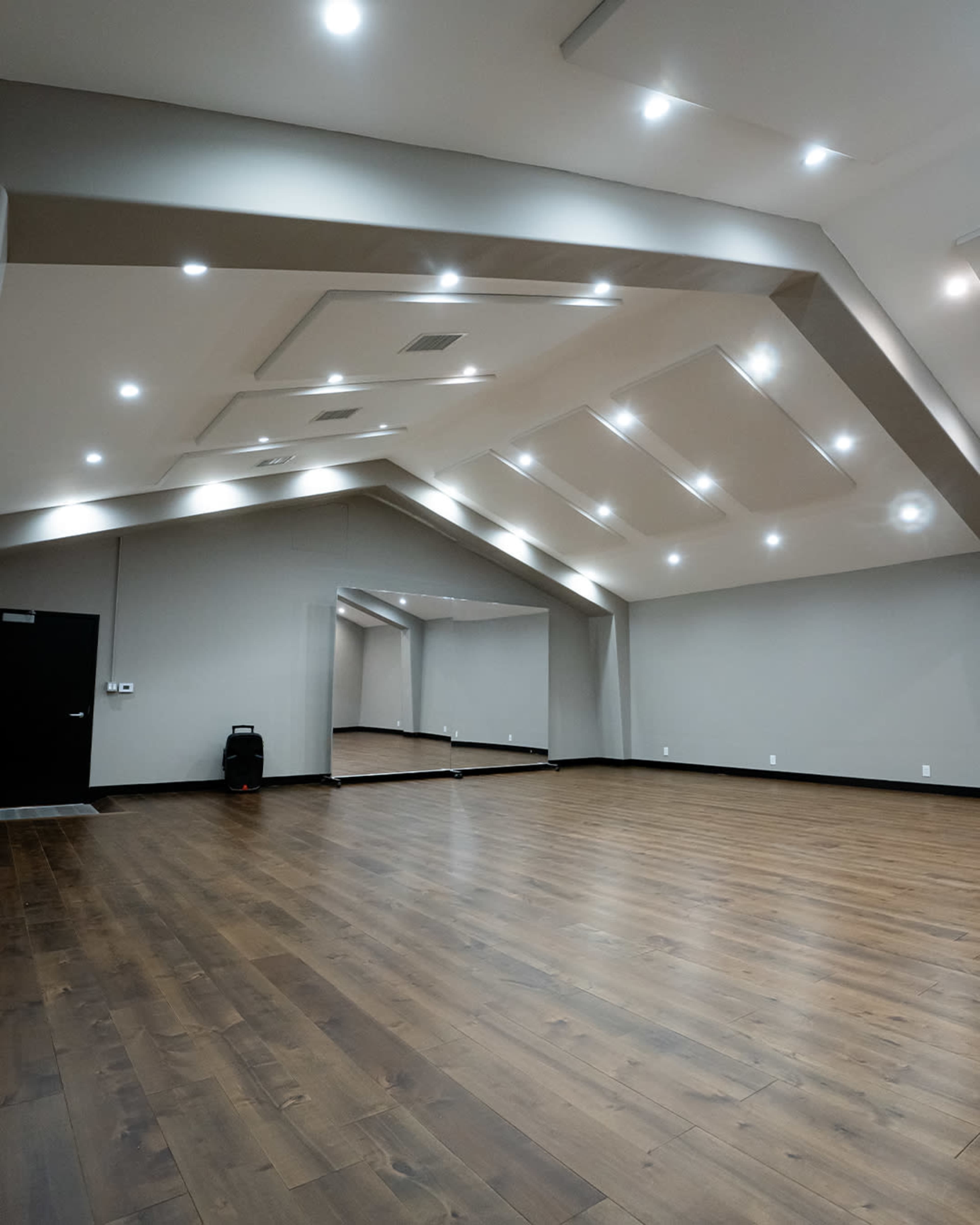 The image shows an empty dance studio with wooden flooring, high ceilings, and wall mirrors illuminated by recessed lighting.