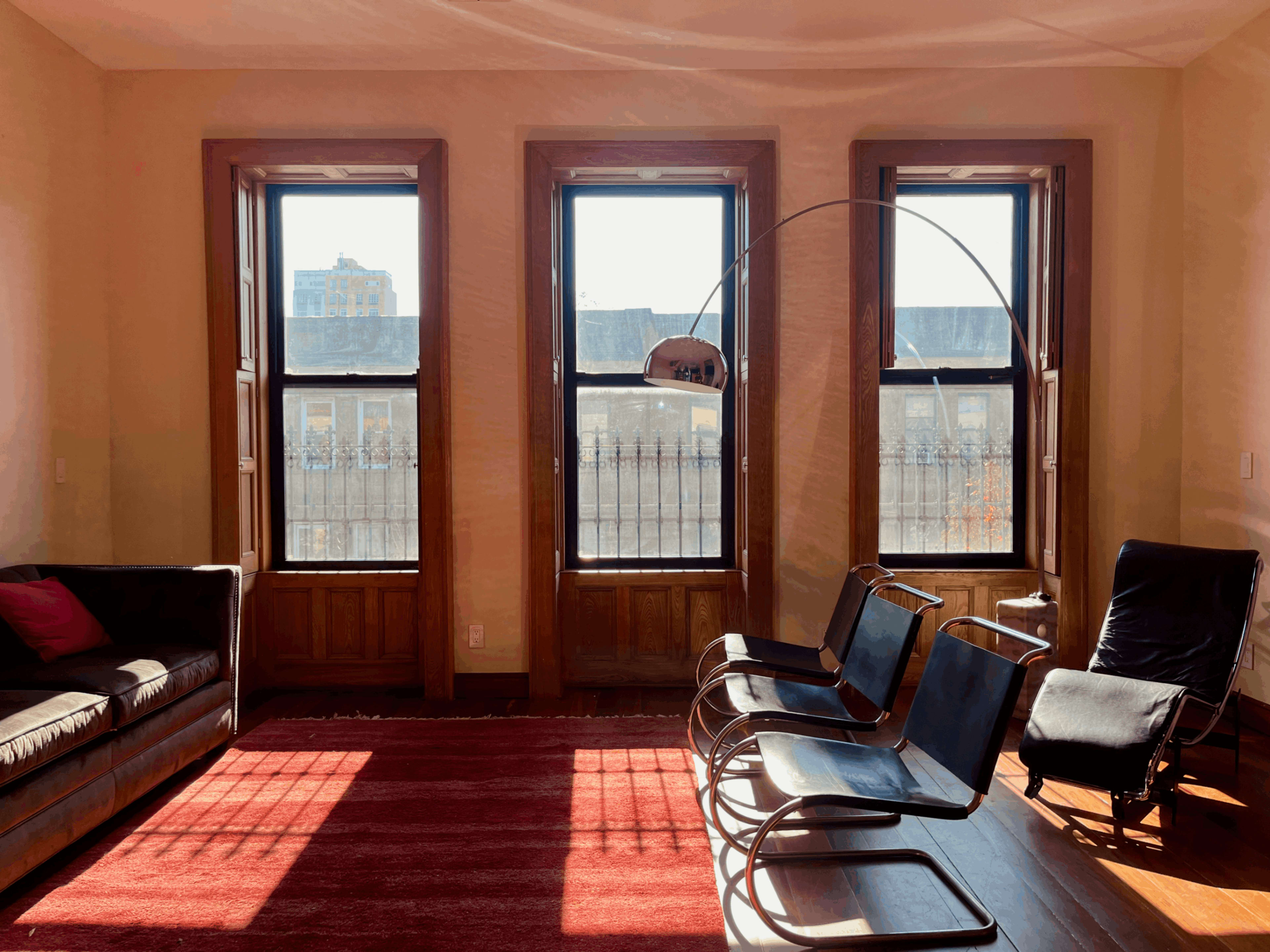 The scene features a sunlit room with three large windows, a dark sofa, two black chairs, and a red rug on the wooden floor.