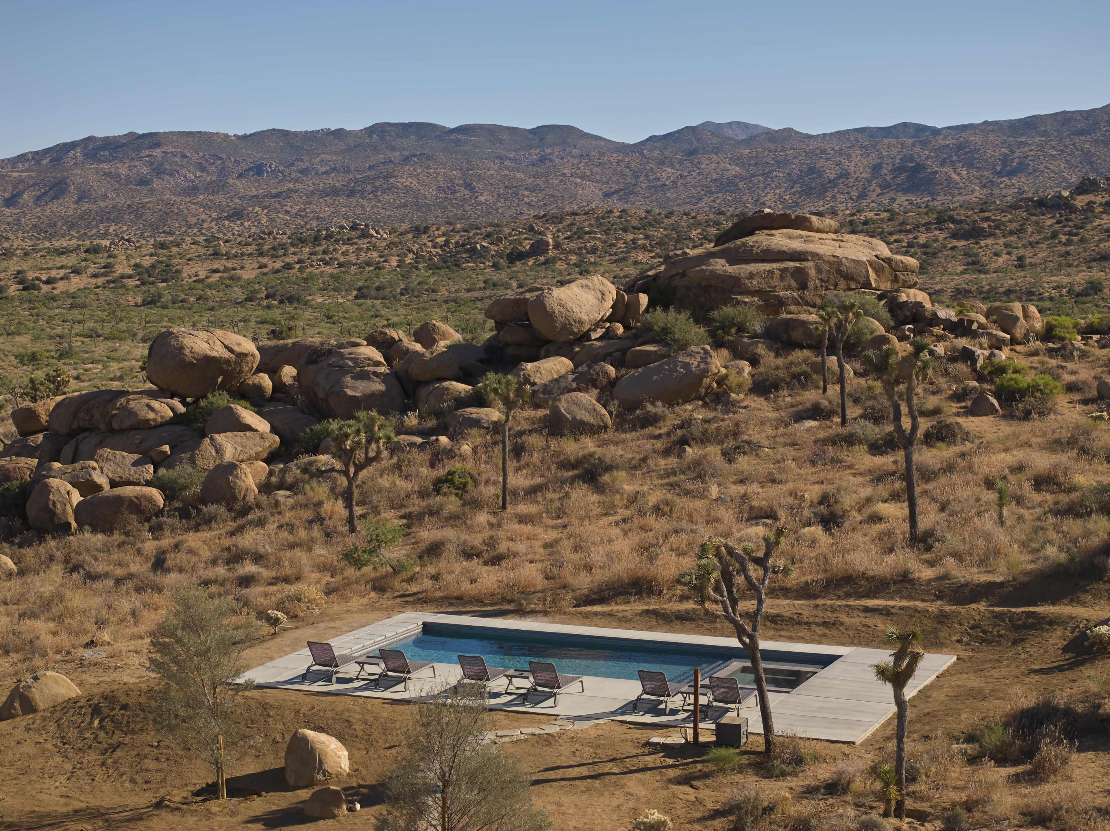 The image shows a swimming pool surrounded by lounge chairs in a desert landscape with rocky hills in the background.