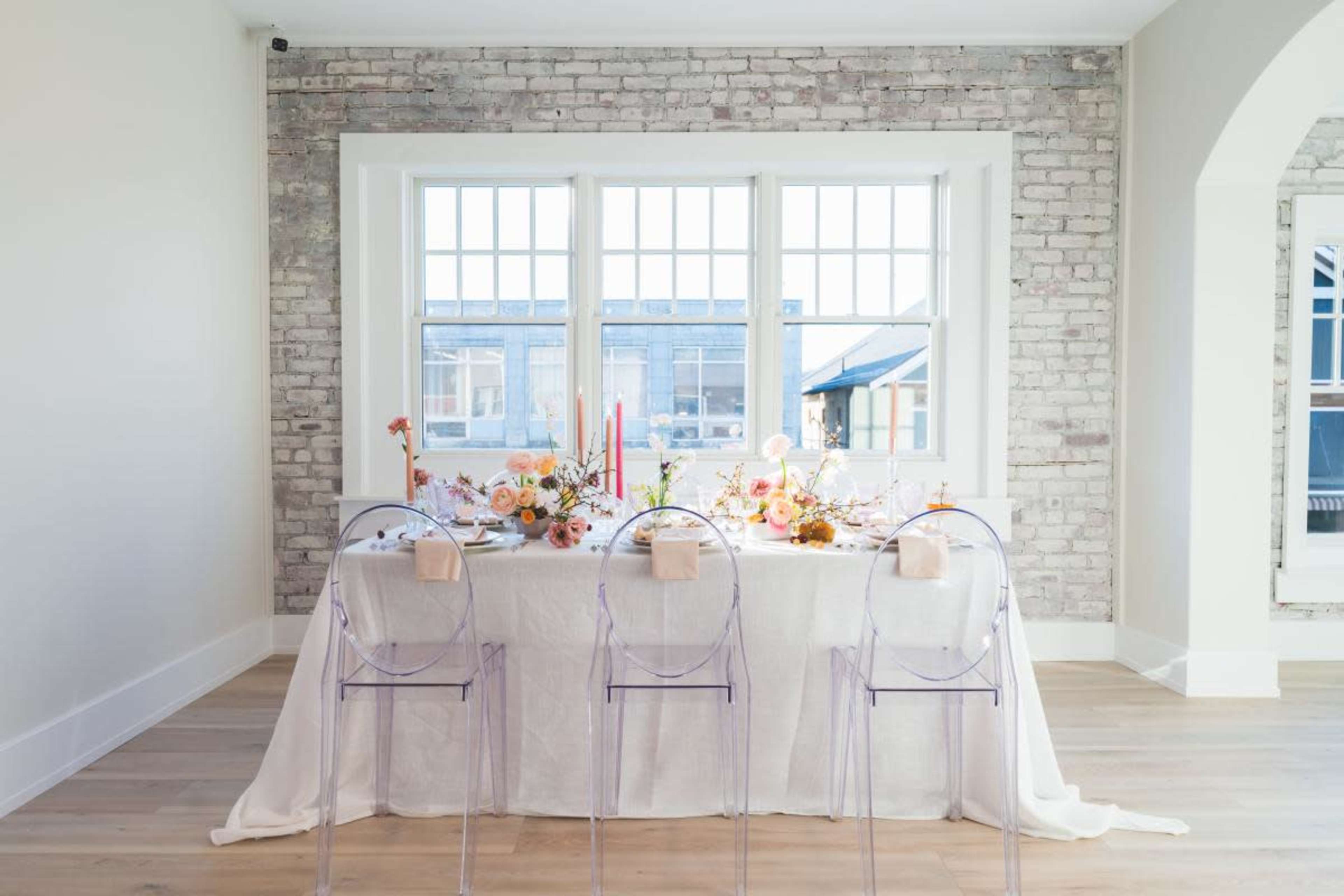 A dining table is elegantly set with flowers, candles, and tableware in front of large windows featuring a brick wall backdrop.
