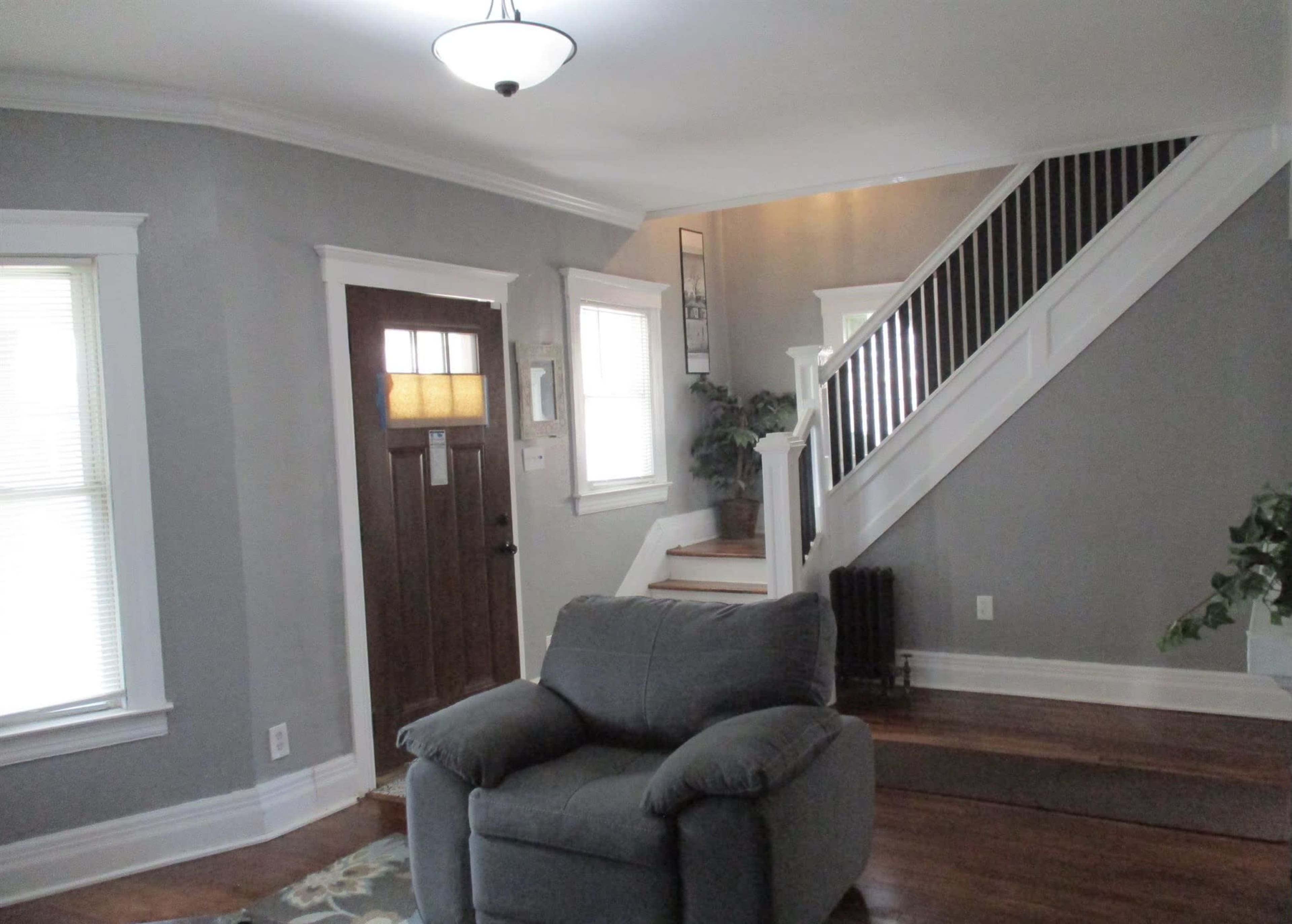The image shows a living room with a gray wall, a brown front door, a staircase in the background, and a gray armchair in the foreground.