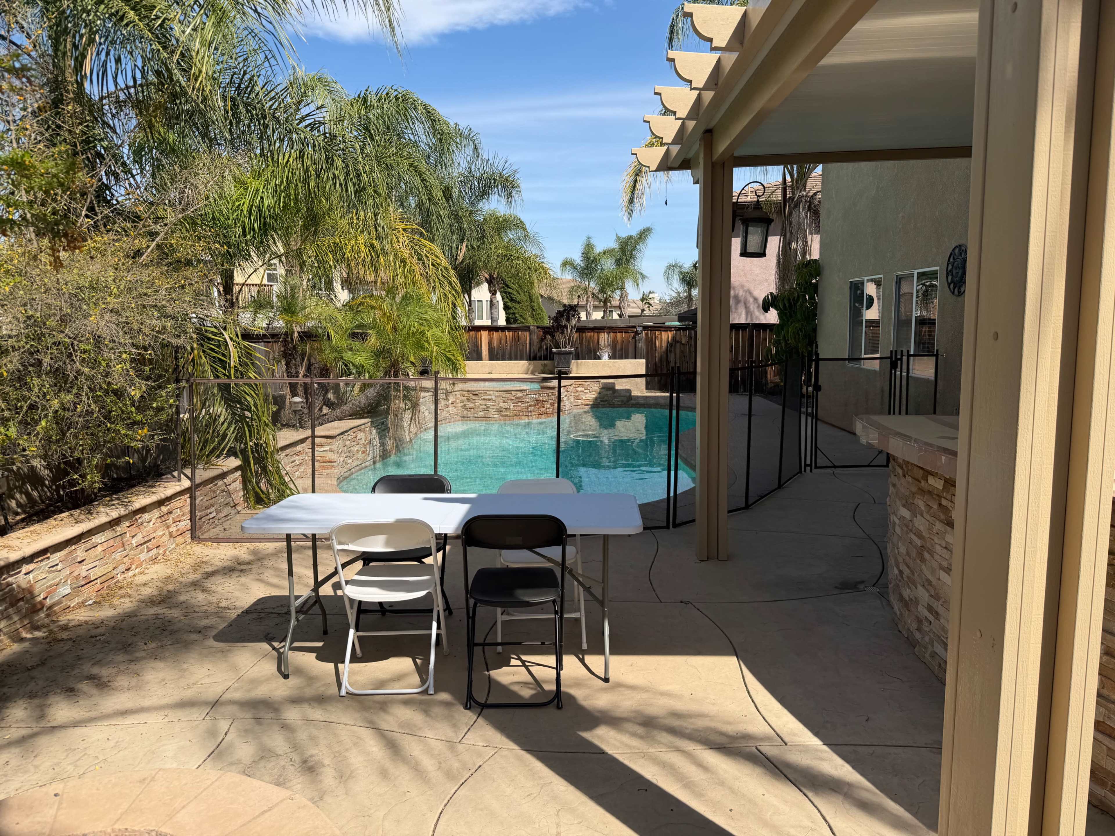 The image shows a backyard with a swimming pool surrounded by palm trees, a seating area with a table and chairs, and a house in the background.