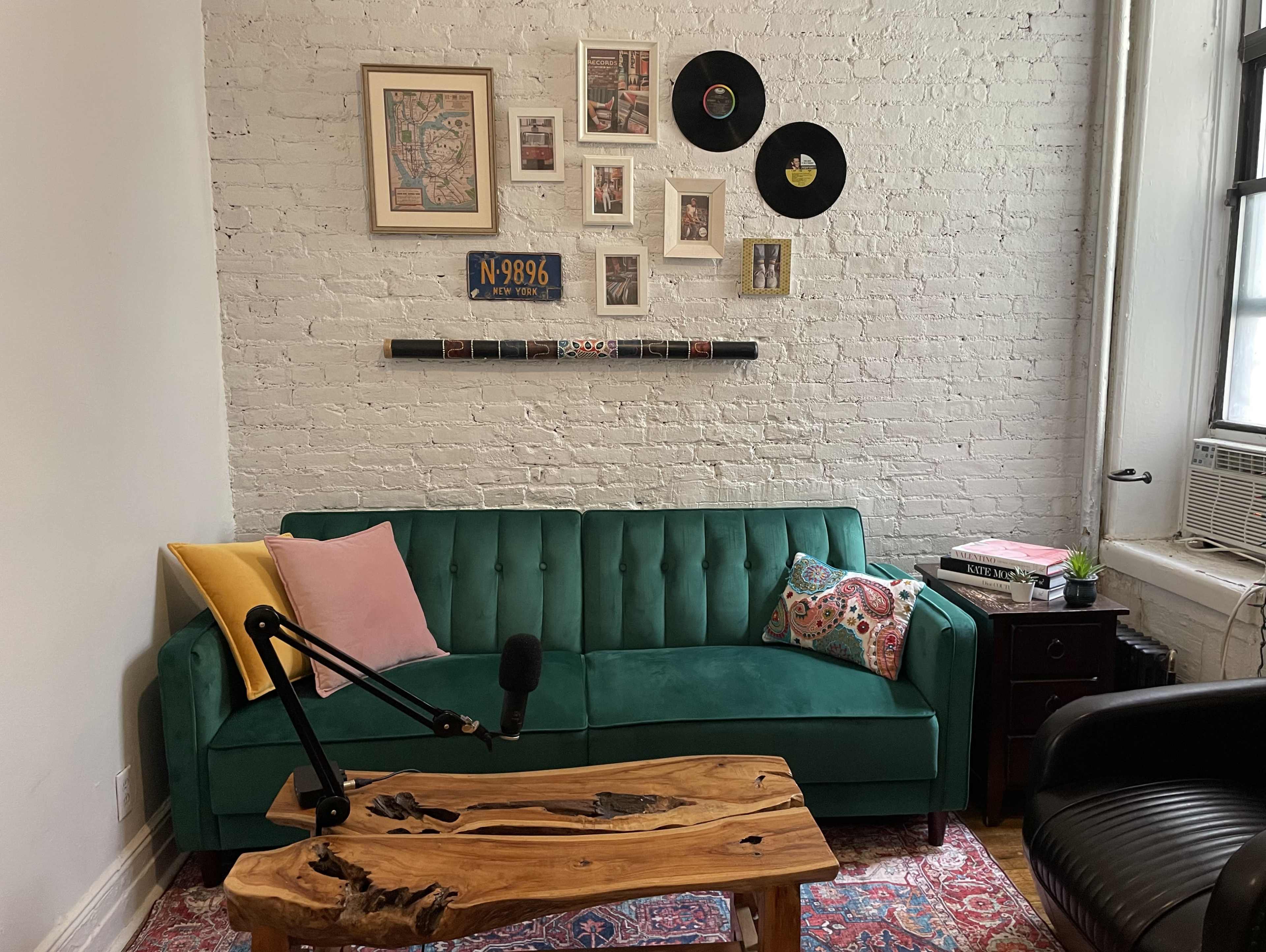 A green couch with decorative pillows against a white brick wall, featuring a gallery of framed art and vinyl records above it, along with a wooden coffee table and a black chair.