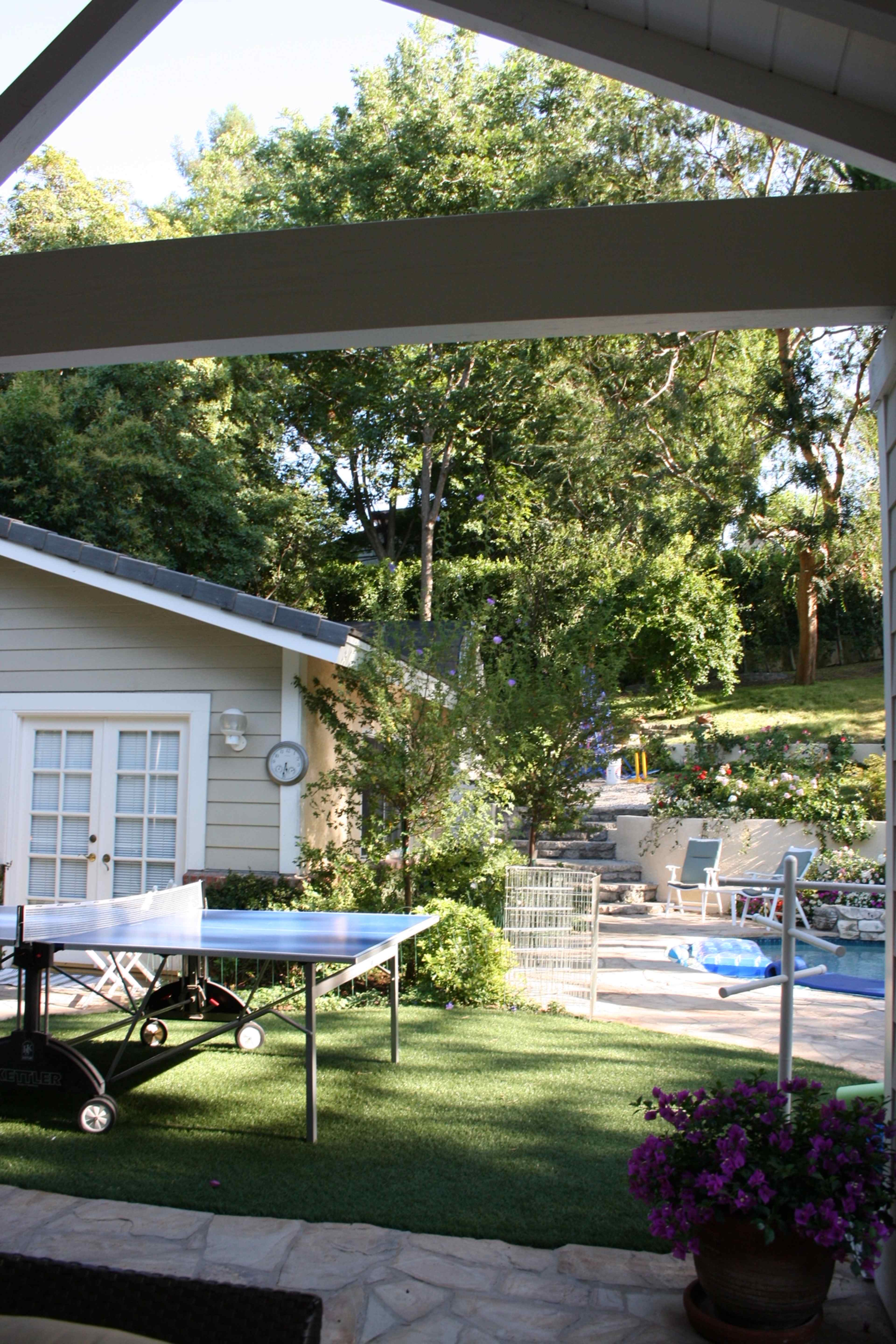 A backyard features a ping pong table on artificial grass, with a pool and garden area in the background.