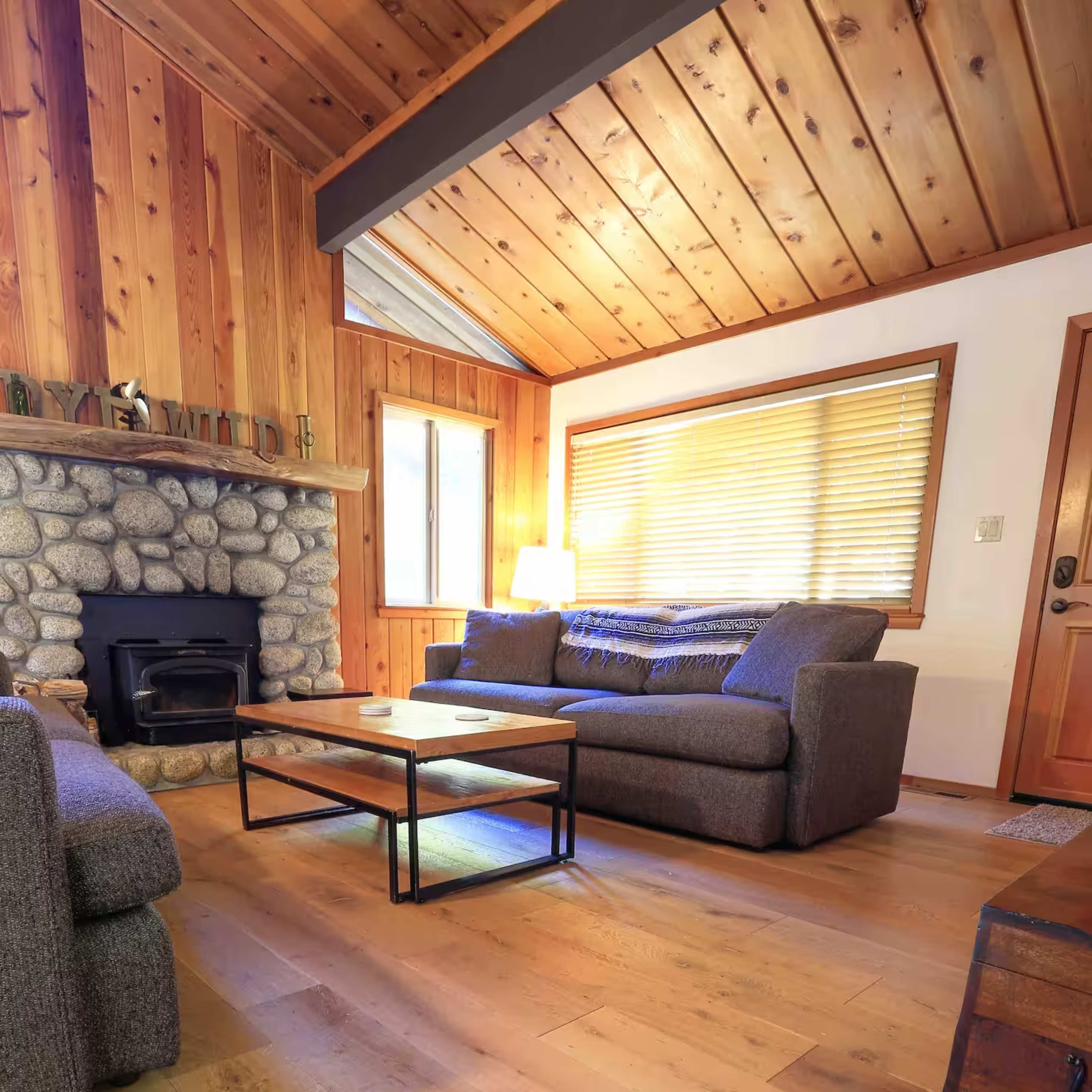 The image shows a cozy living room with a stone fireplace, wooden walls, and a simple coffee table between two gray sofas.