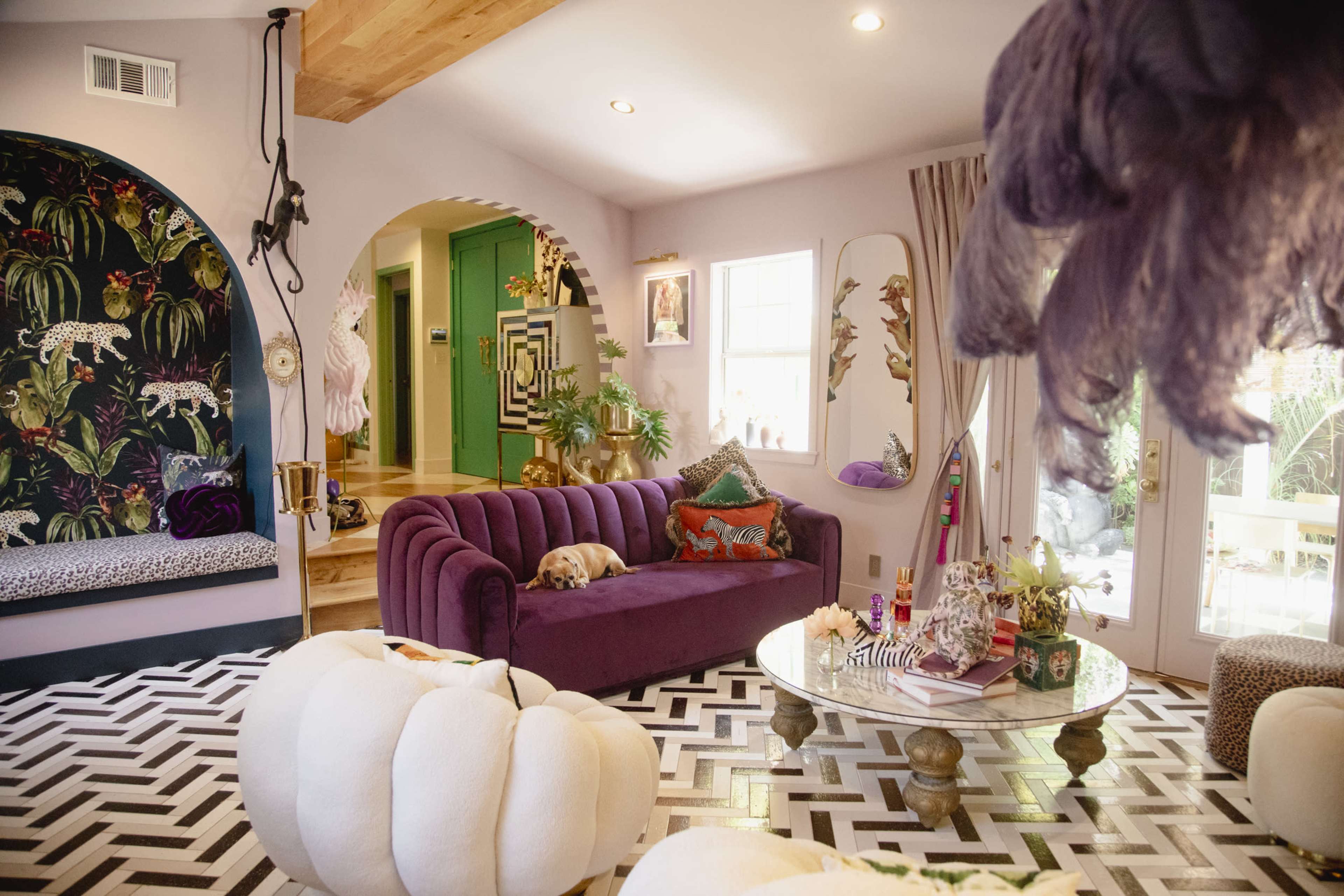 The image shows a brightly decorated living room with a purple sofa, a geometric black-and-white patterned floor, and colorful decor elements on various surfaces.