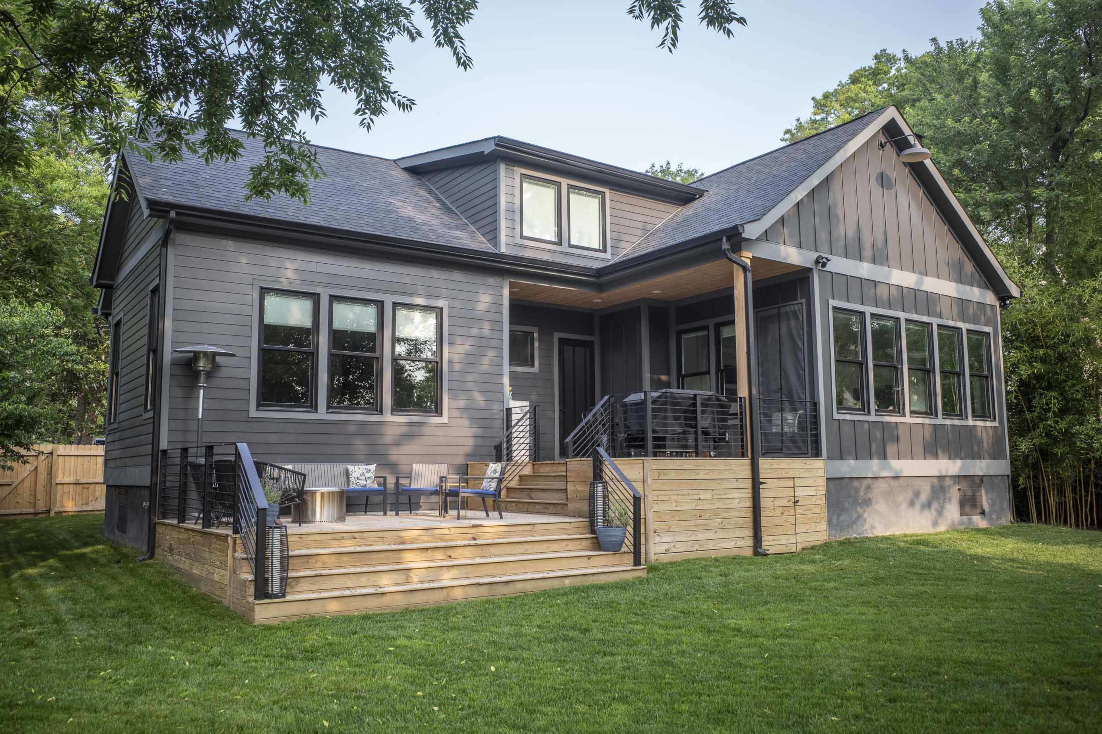 A modern single-story house with dark siding and a spacious wooden deck is surrounded by green grass and trees.