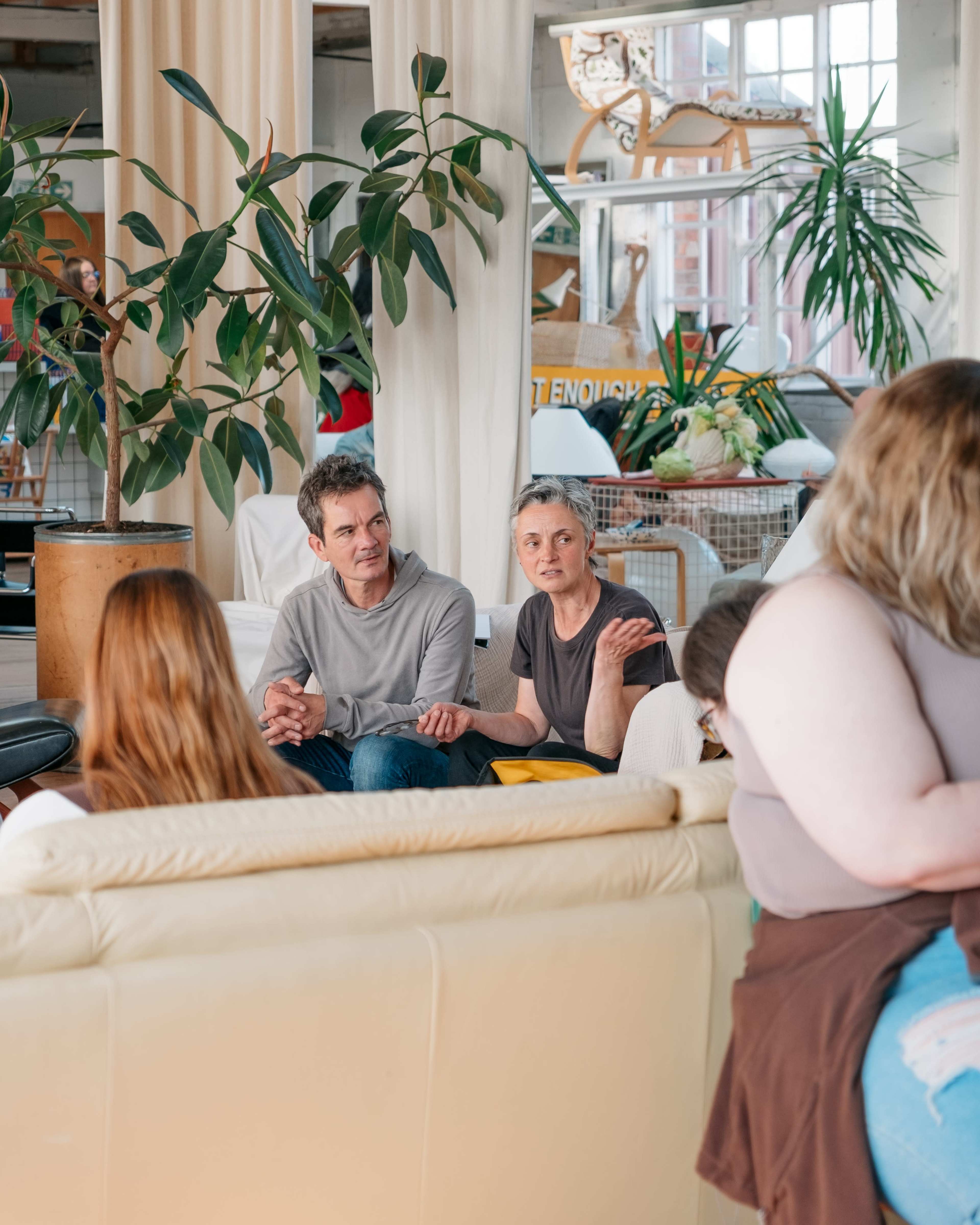 A man and a woman sit together on a white couch in a bright, modern interior with plants and light-colored curtains.