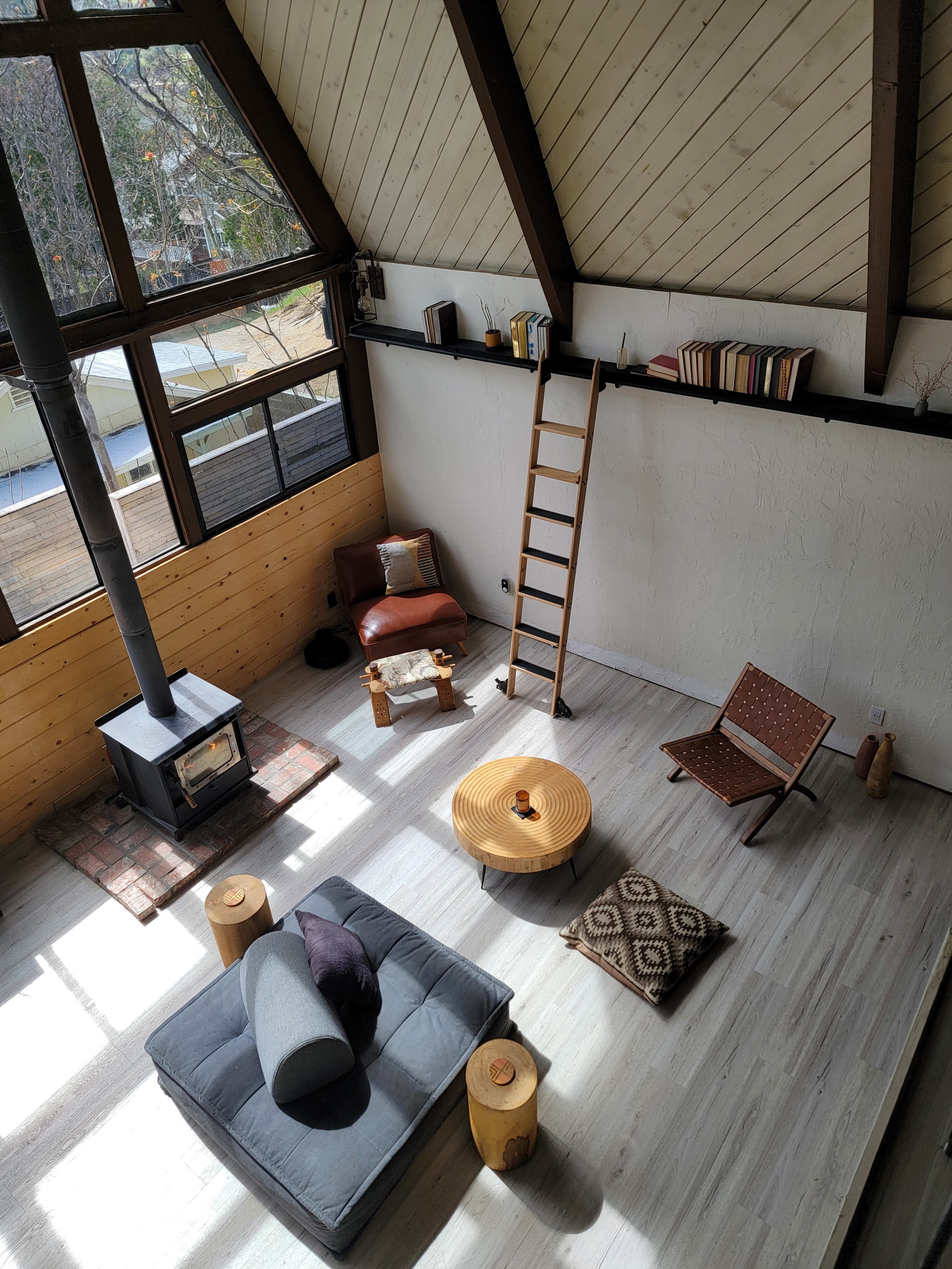 A modern, open living space with a wood stove, a ladder leading to a loft, and various seating arrangements including a floor cushion and a wooden table.