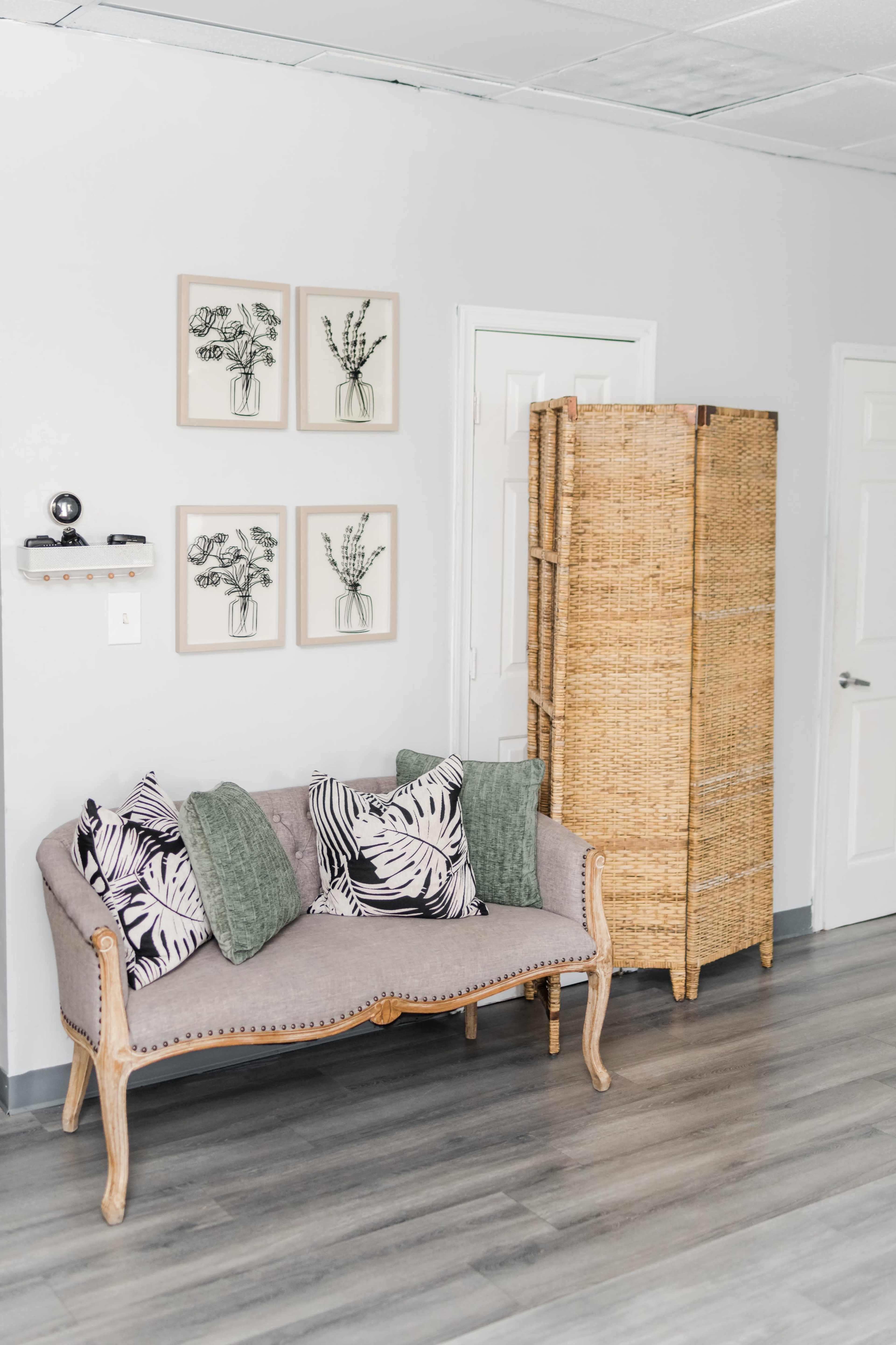 A light gray sofa with patterned pillows is positioned near a woven room divider and framed botanical prints on the wall.