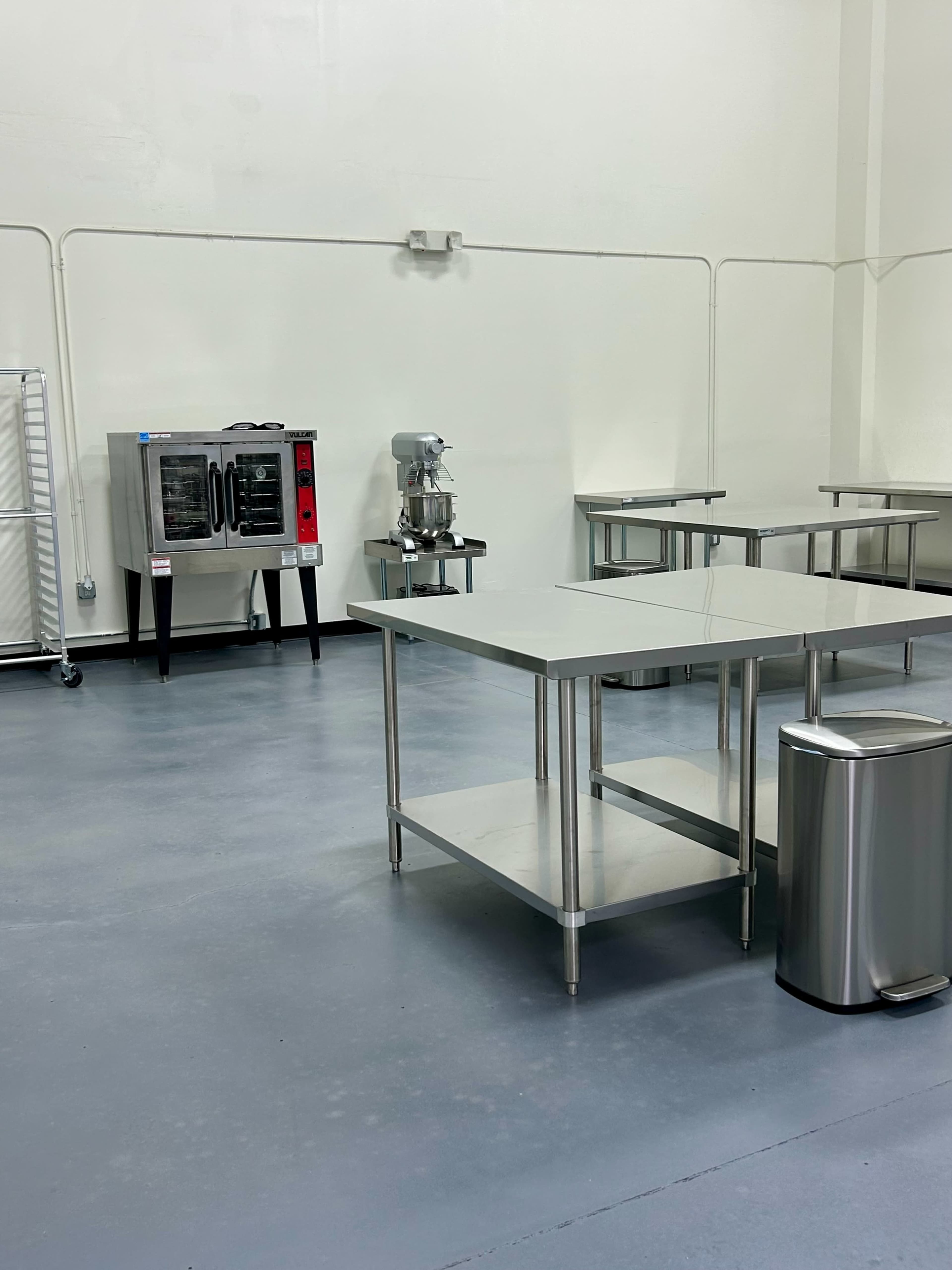 The image shows a clean, empty commercial kitchen space with stainless steel tables, a cooking oven, a mixer, and a trash can.