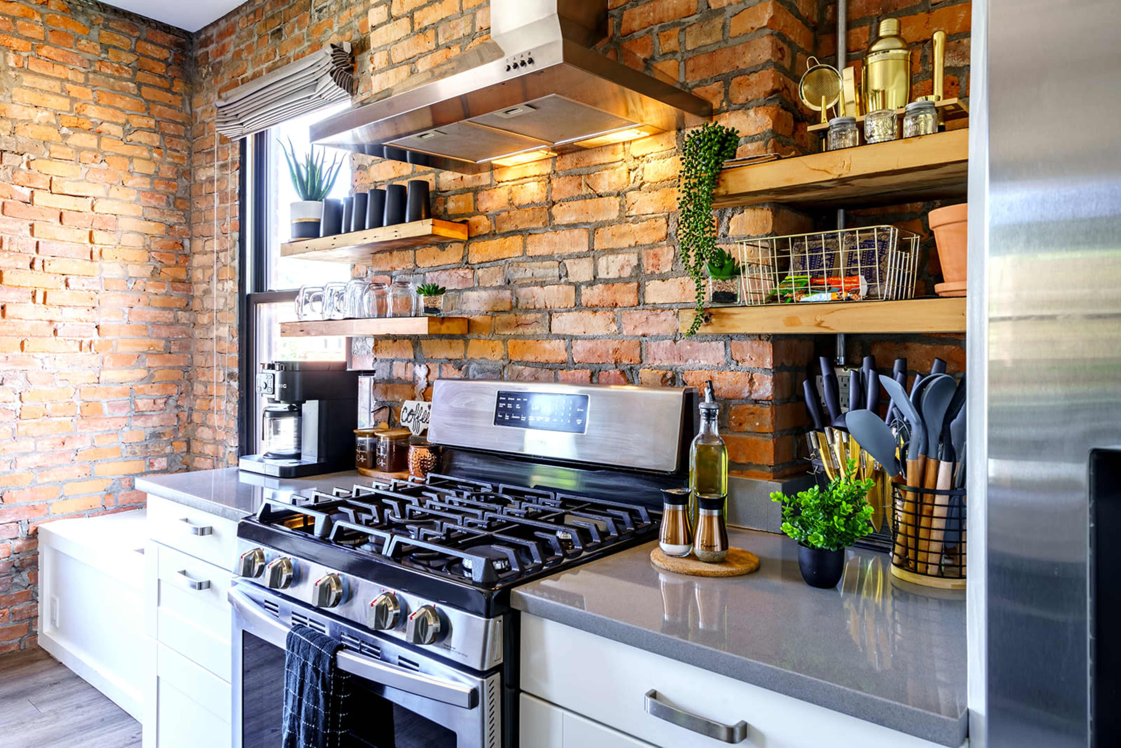 A modern kitchen features a stainless steel gas stove, gray countertops, wooden shelves with various plants and kitchen items, and exposed brick walls.