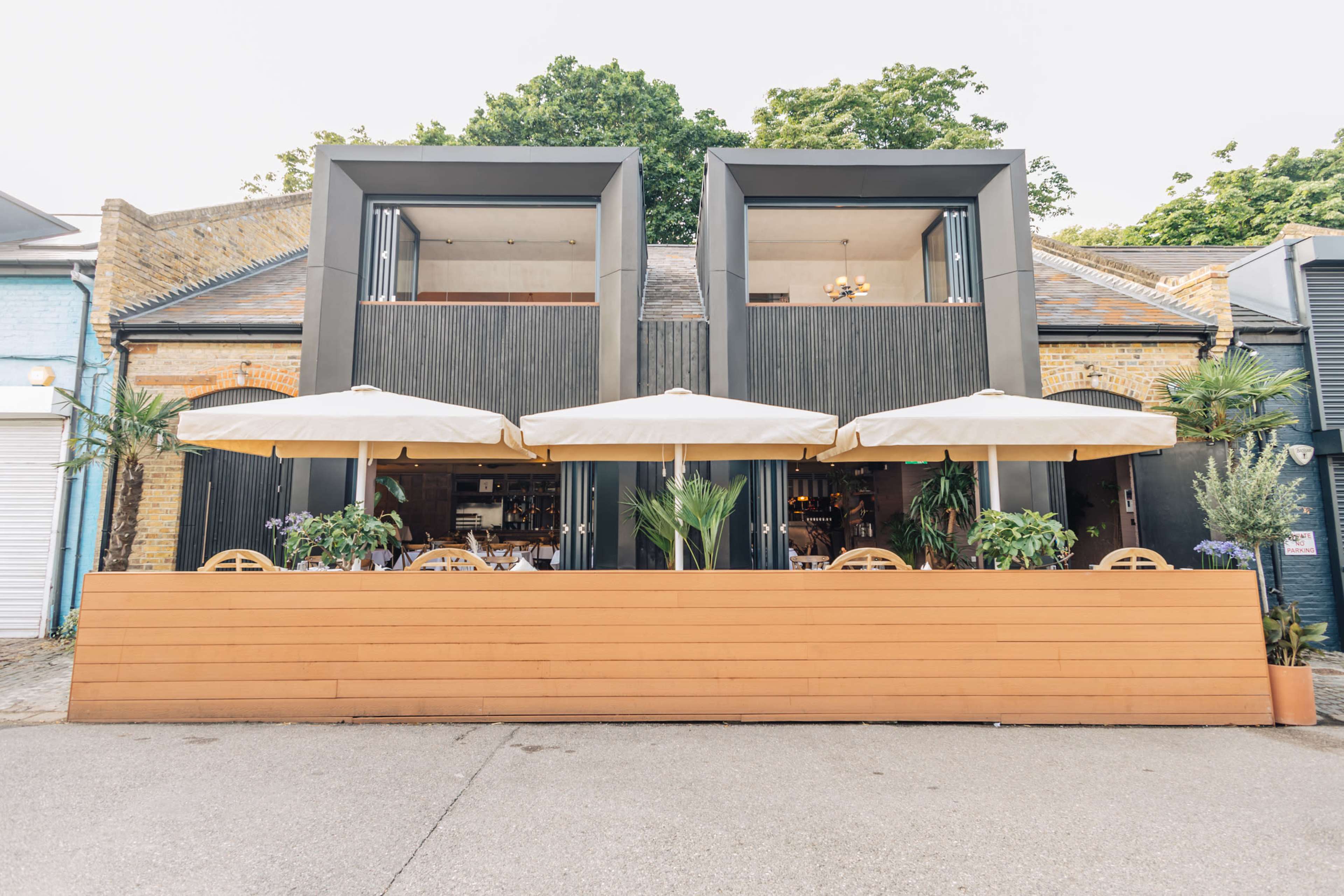 The image shows a two-story building with large, square windows, outdoor seating under umbrellas, and planters at the front.