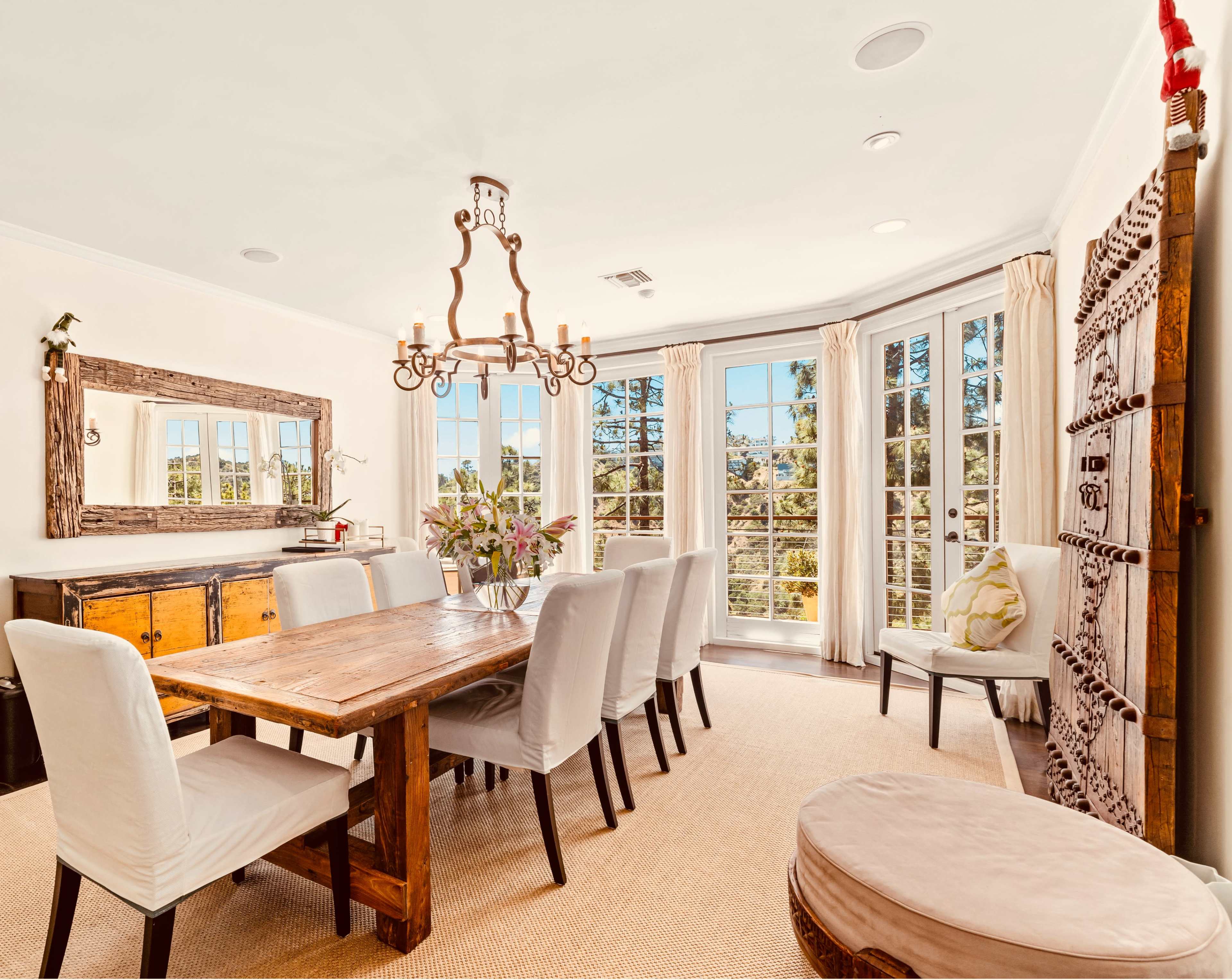 A dining room features a large wooden table surrounded by upholstered chairs, with a chandelier overhead and large windows allowing natural light.