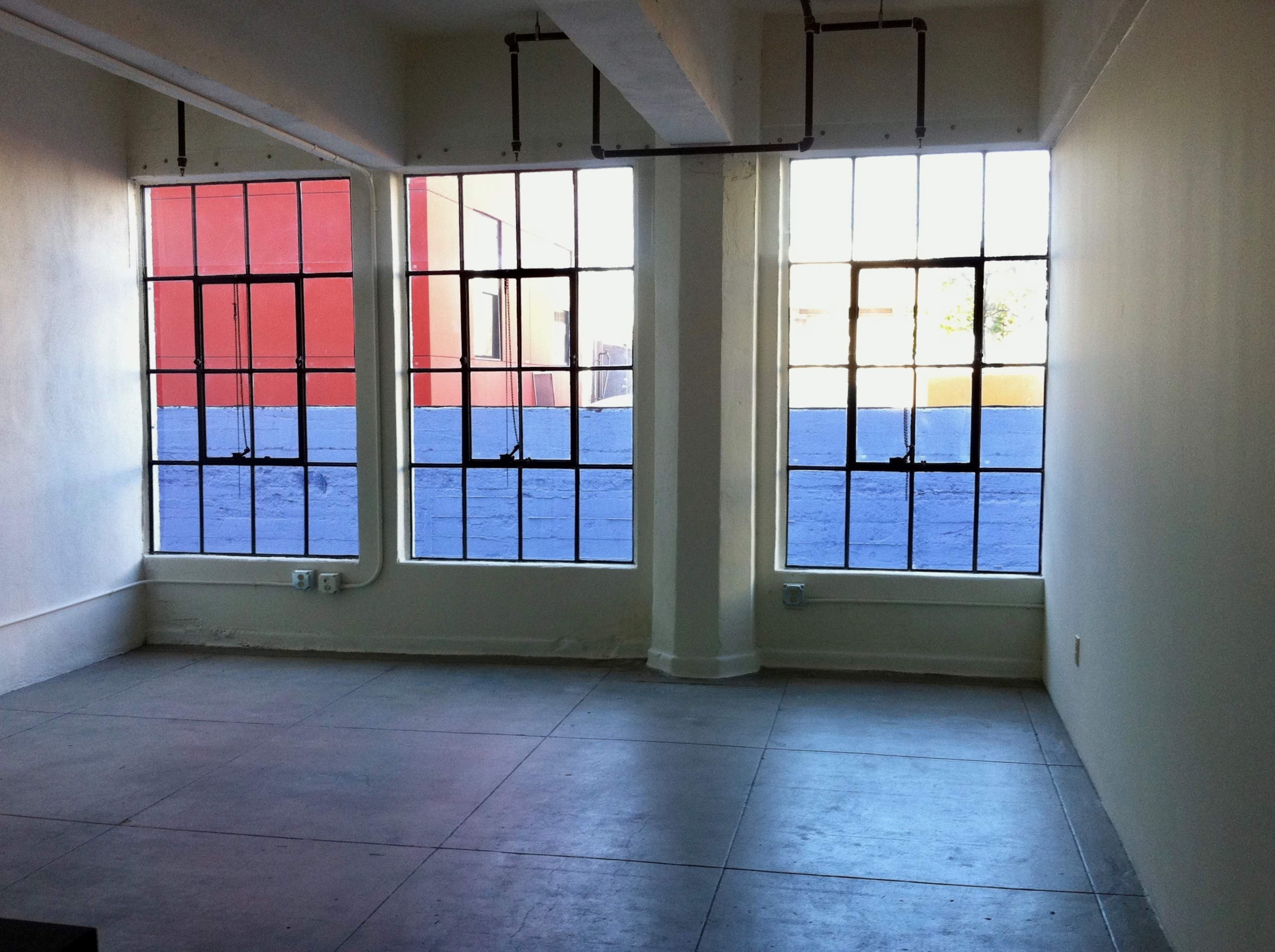 The image depicts an empty room with large, industrial-style windows that have black frames, allowing light to enter and reveal a concrete floor.
