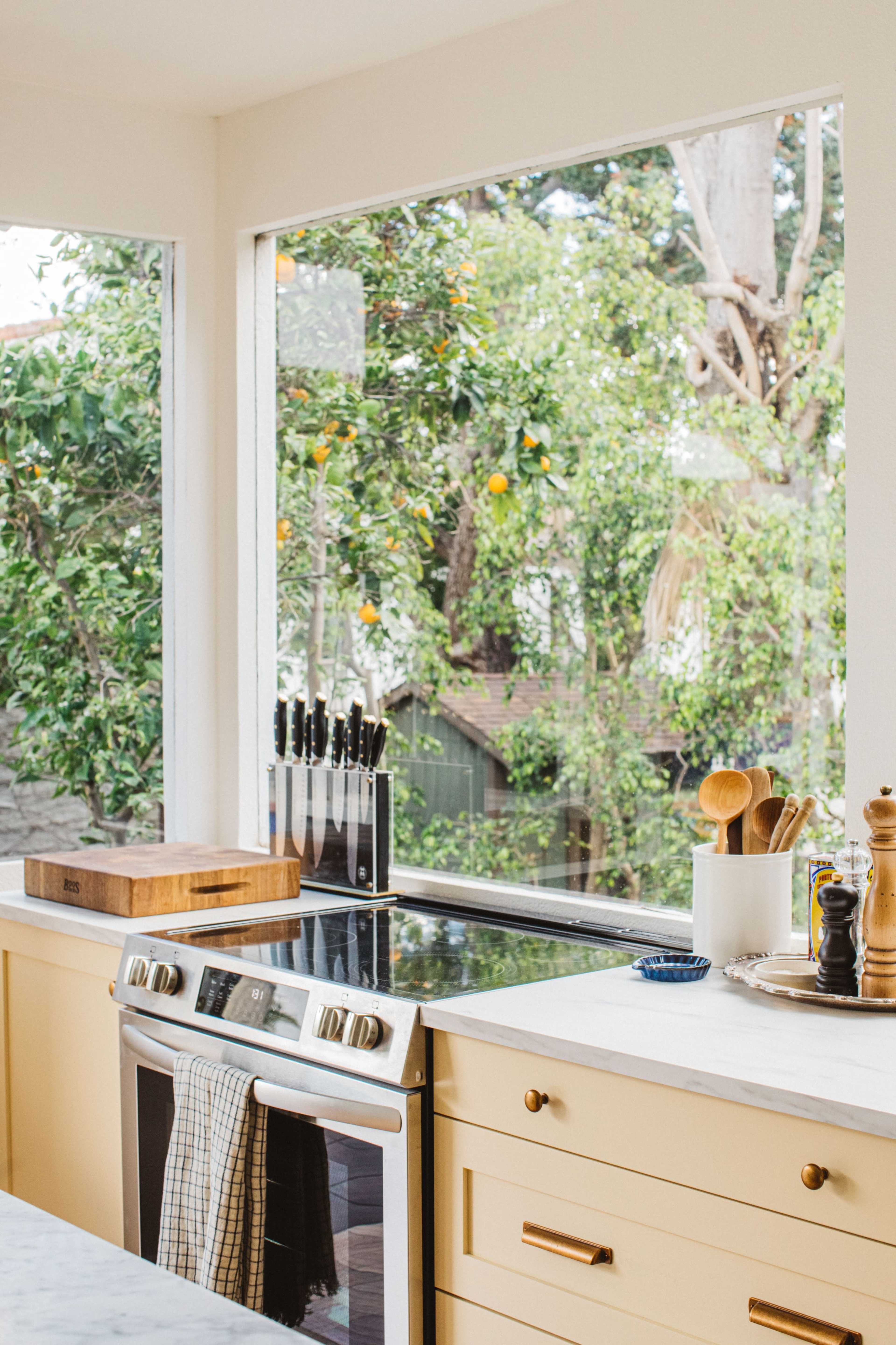 A modern kitchen features a stovetop, a wooden cutting board, knife block, and utensils, with large windows showcasing green trees and orange fruit outside.