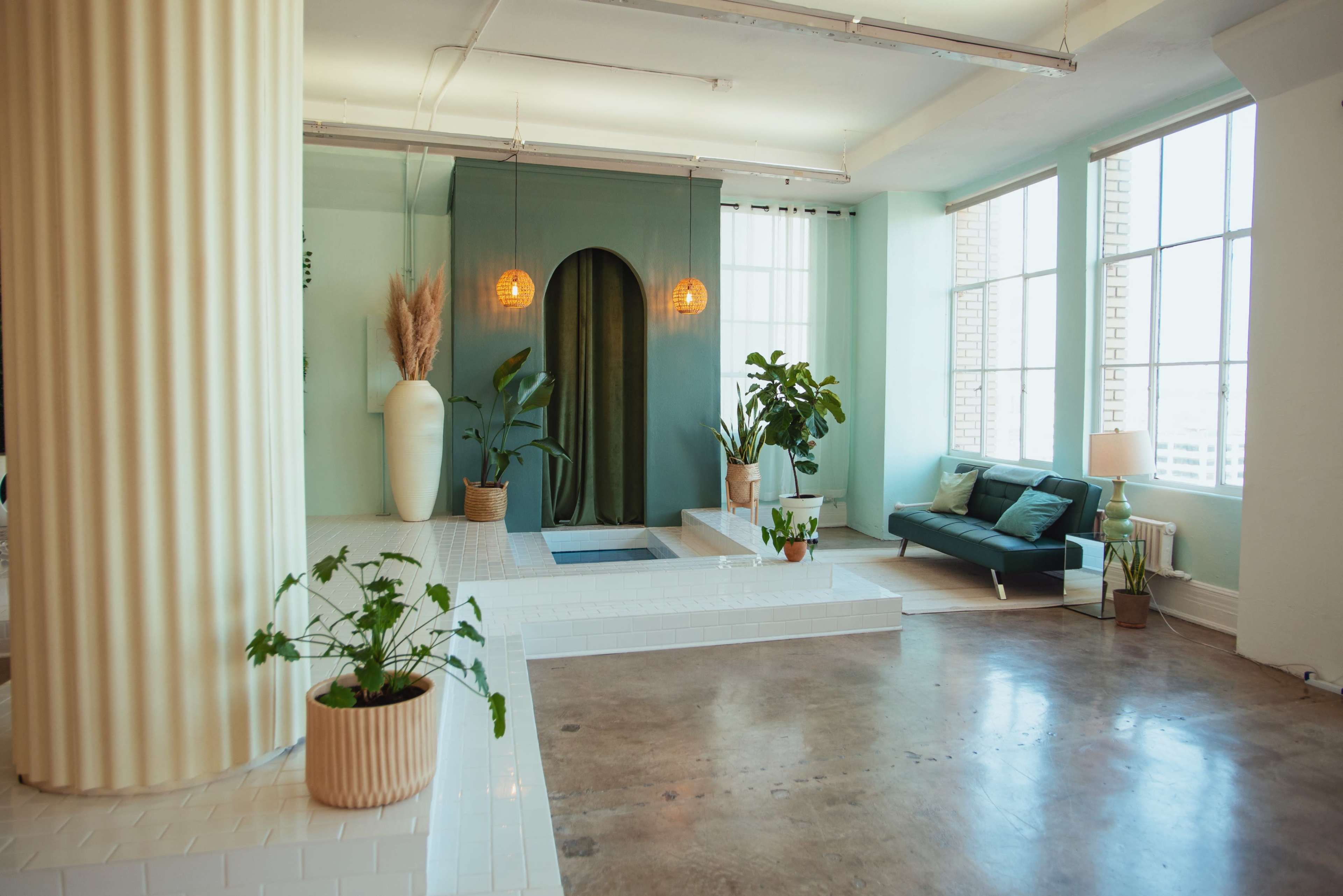 A modern interior space featuring large windows, a minimalist green corner with hanging lights, various plants, and a sofa.