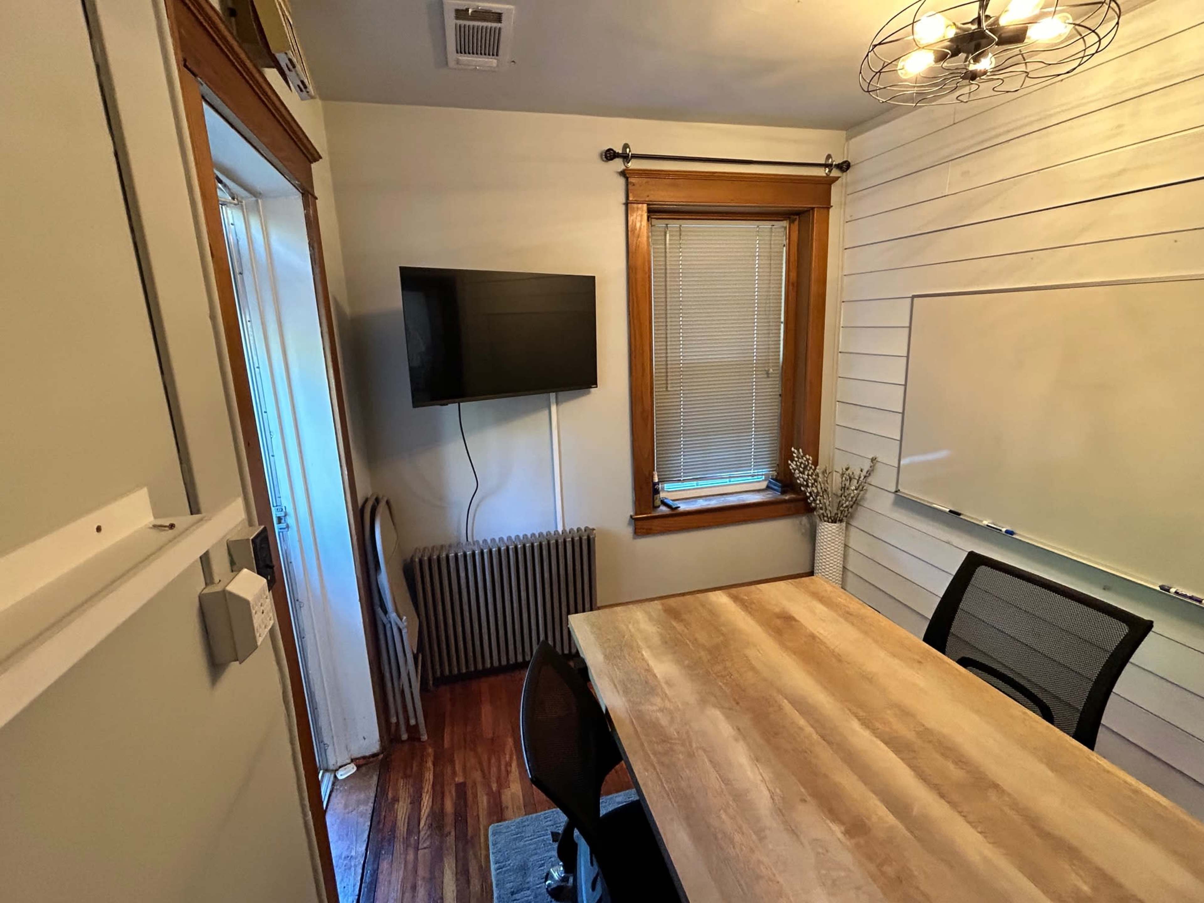 The image shows a small room with a wooden table and chairs, a TV mounted on the wall, and a whiteboard next to a window.