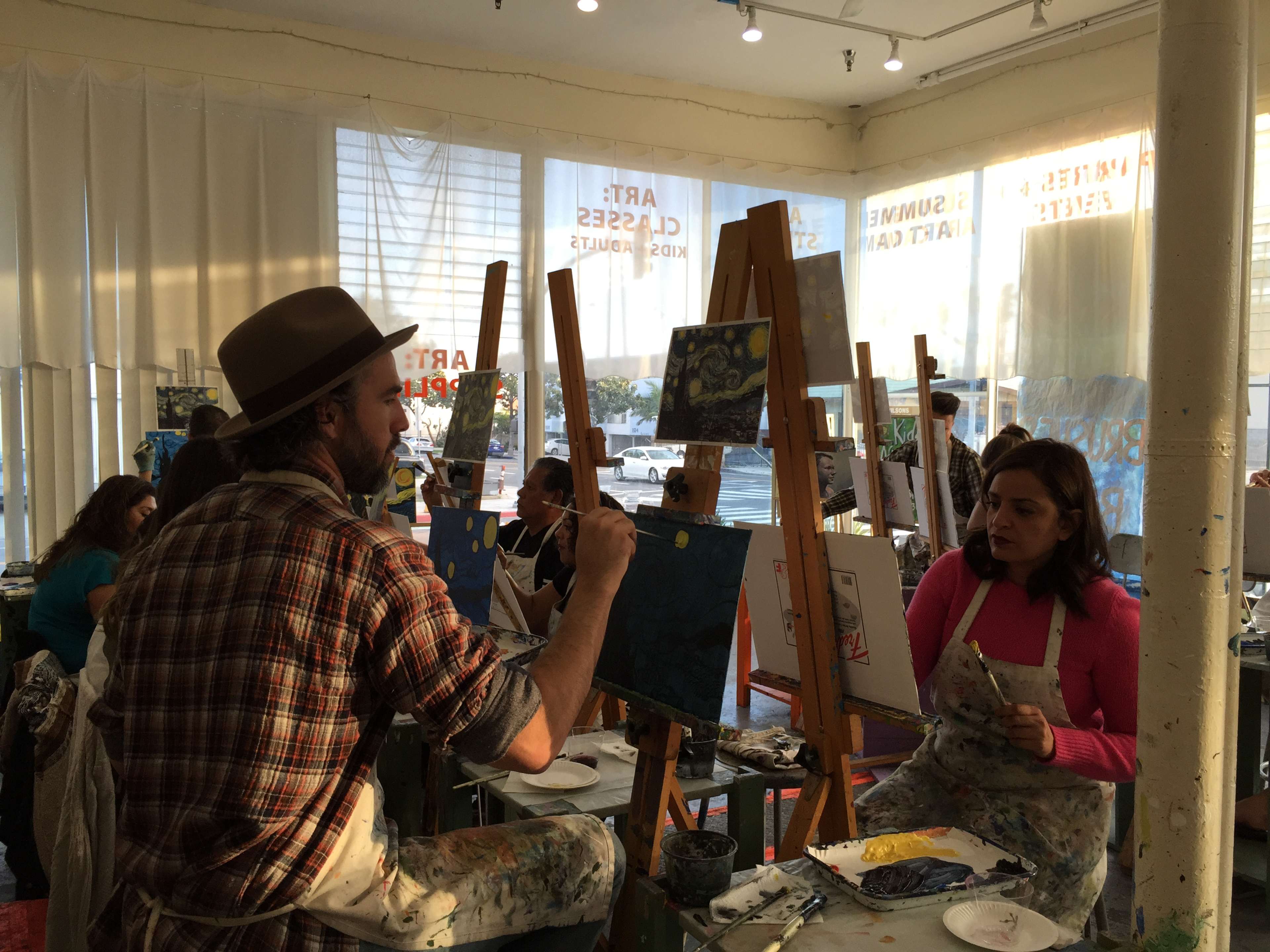 A group of artists paints on canvases in a sunlit studio with large windows.